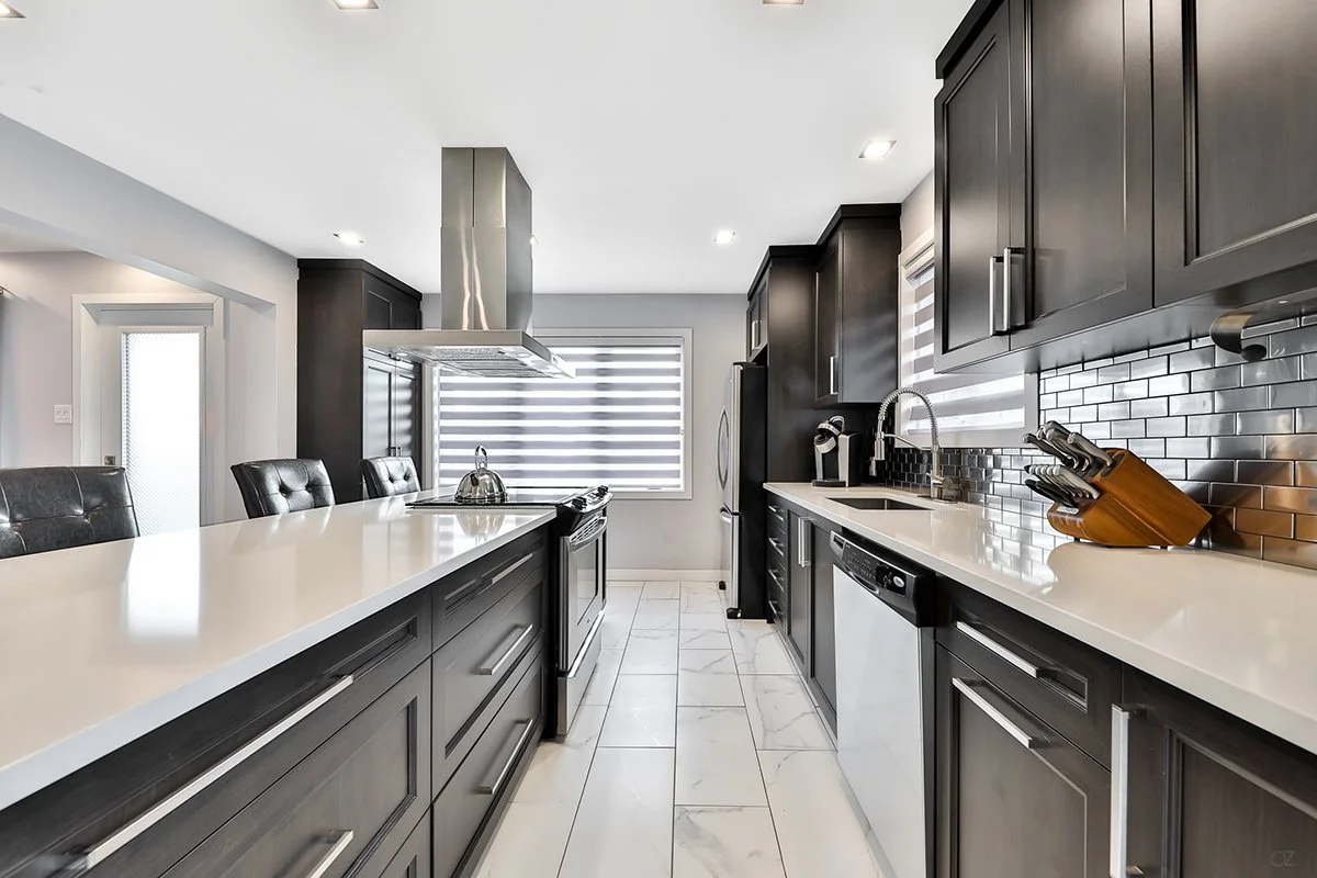 Cuisine moderne avec comptoir en quartz blanc, armoires en bois foncé, appareils en acier inoxydable, avec une fenêtre à stores horizontaux