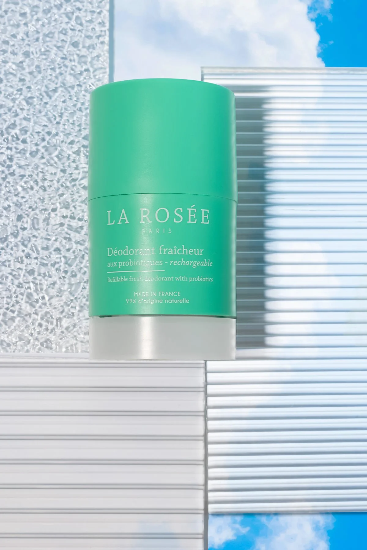 Déodorant en spray vert avec texte en français et anglais, sous un ciel bleu, posé sur une surface blanche en plastique à lames.