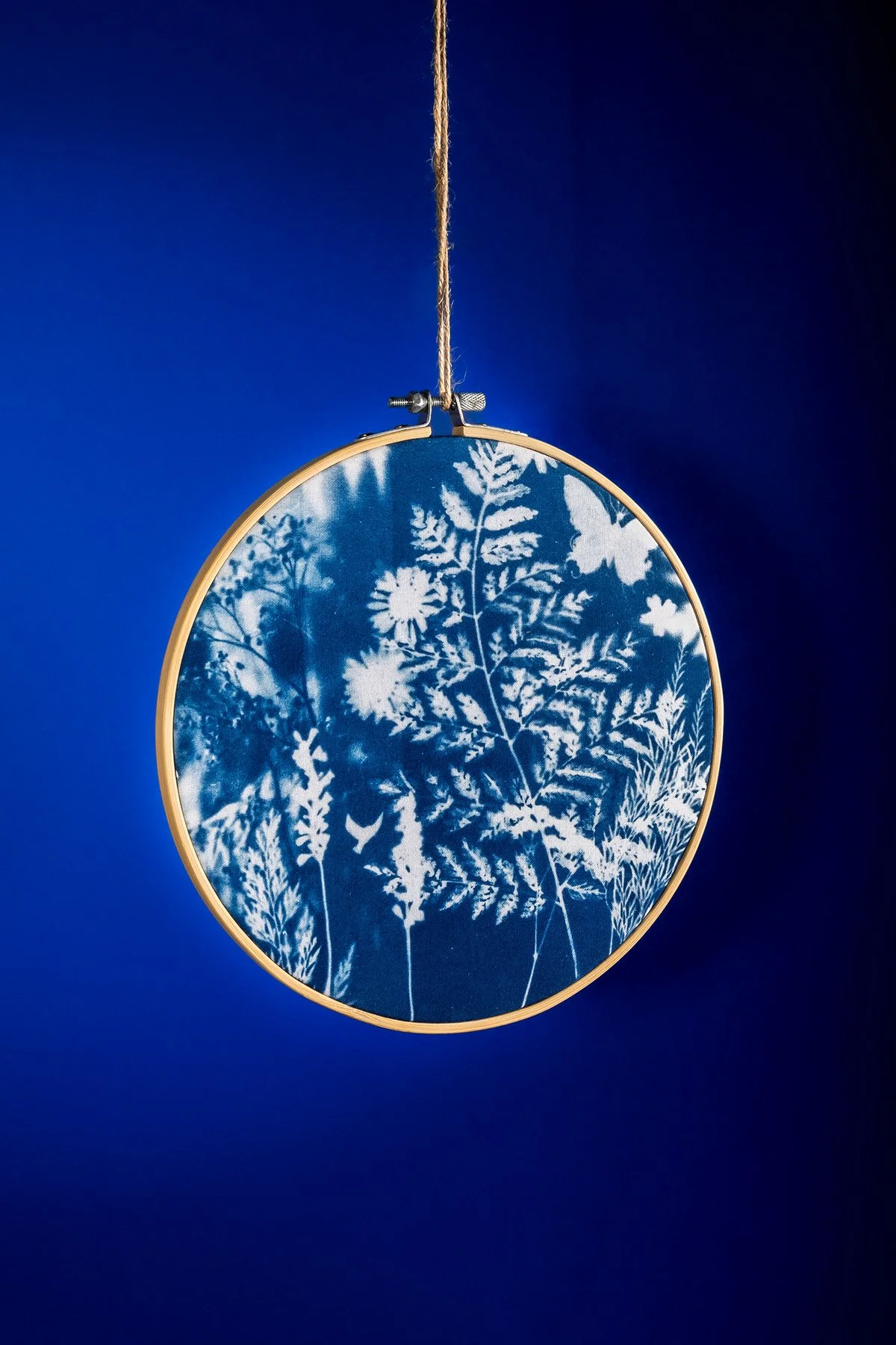 Une œuvre d'art suspendue représentant des silhouettes d'herbes et de fleurs blanches sur un fond bleu, encadrée d'un cercle en bois, suspendue par une corde sur un fond bleu foncé.