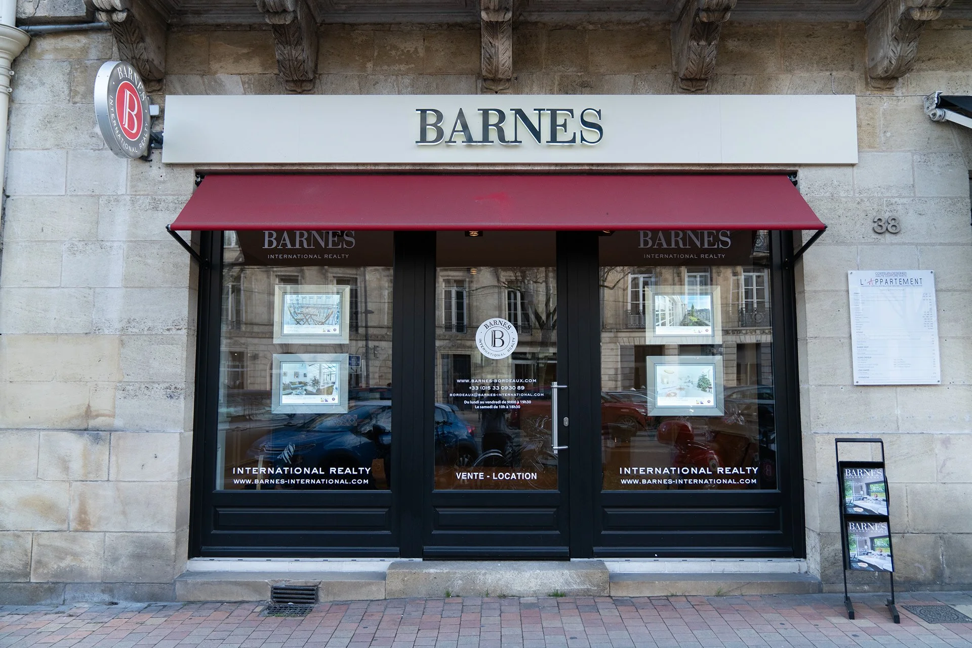 Vitrine d'agence immobilière "Barnes" avec enseigne en lettres noires, au-dessus d'une porte en verre, sous une marquise rouge. Affiches et informations immobilières visibles dans la vitrine.