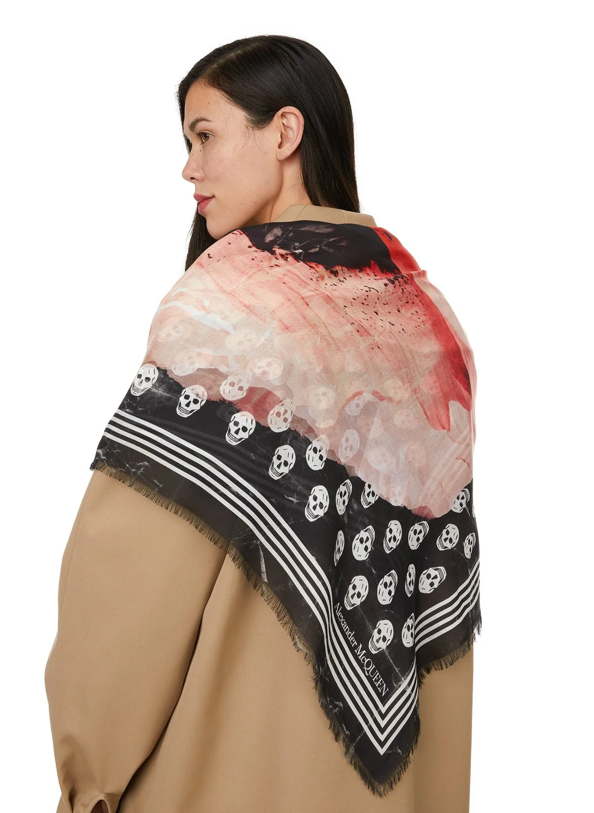 Une femme porte un foulard avec un motif de têtes de mort, des bandes noires et blanches, et le nom 'Alexander McQueen' écrit sur l'angle.