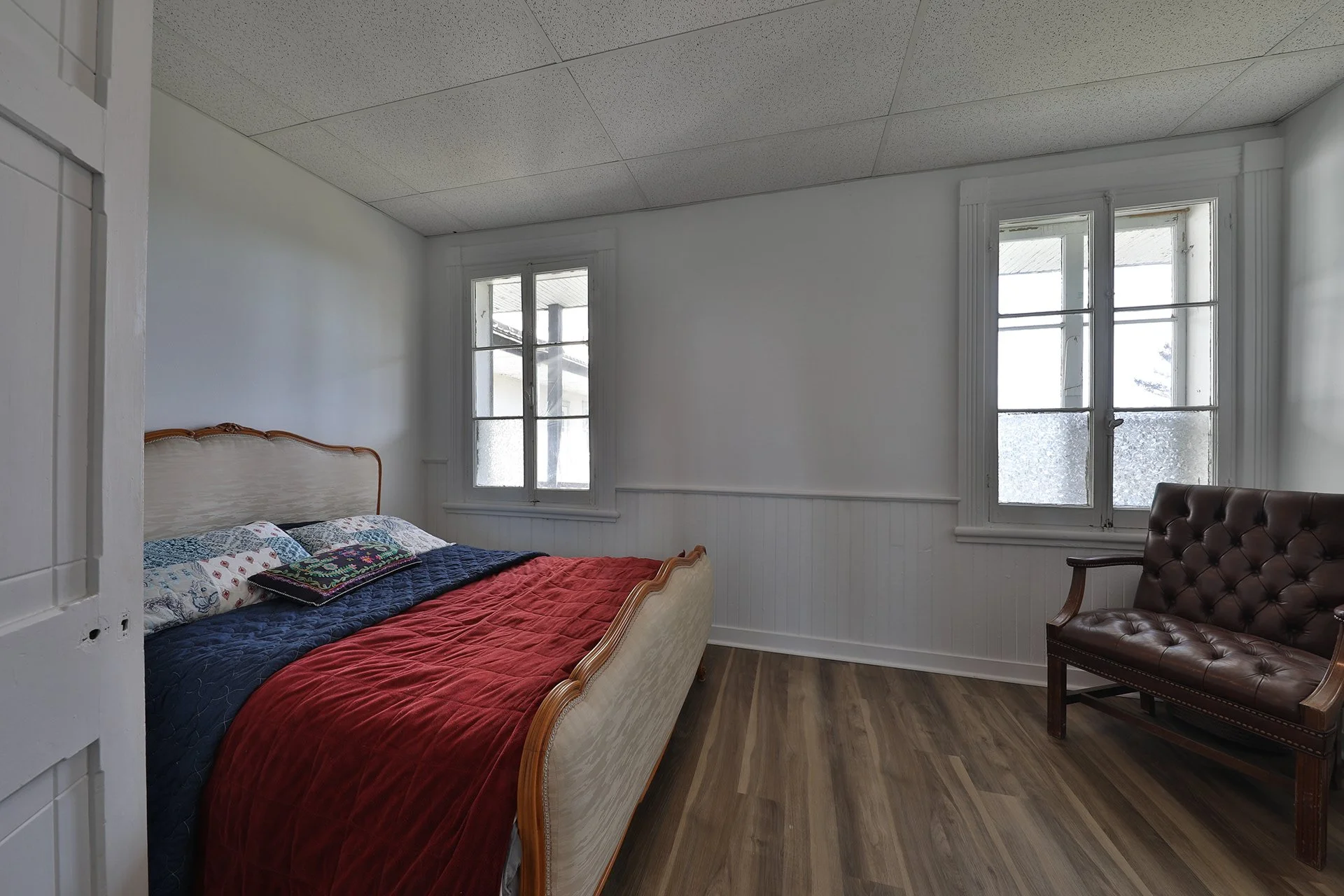 Chambre simple avec lit en bois, couette rouge, oreillers, et fauteuil en cuir marron, deux fenêtres laissant entrer la lumière.