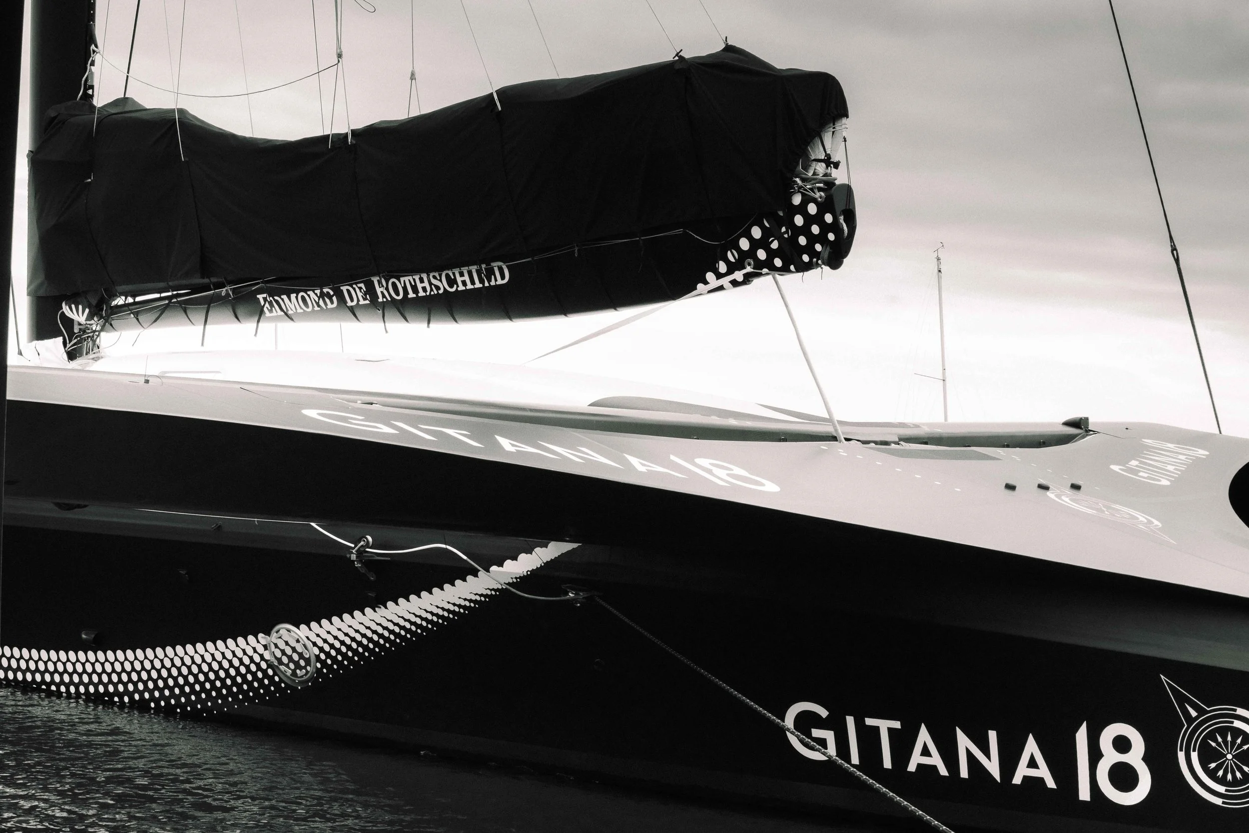 Un voilier nommé GITANA 18 avec une voile noire couverte d'une protection, amarré dans l'eau, avec un logo de la marque Edmond de Rothschild visible sur la voile.