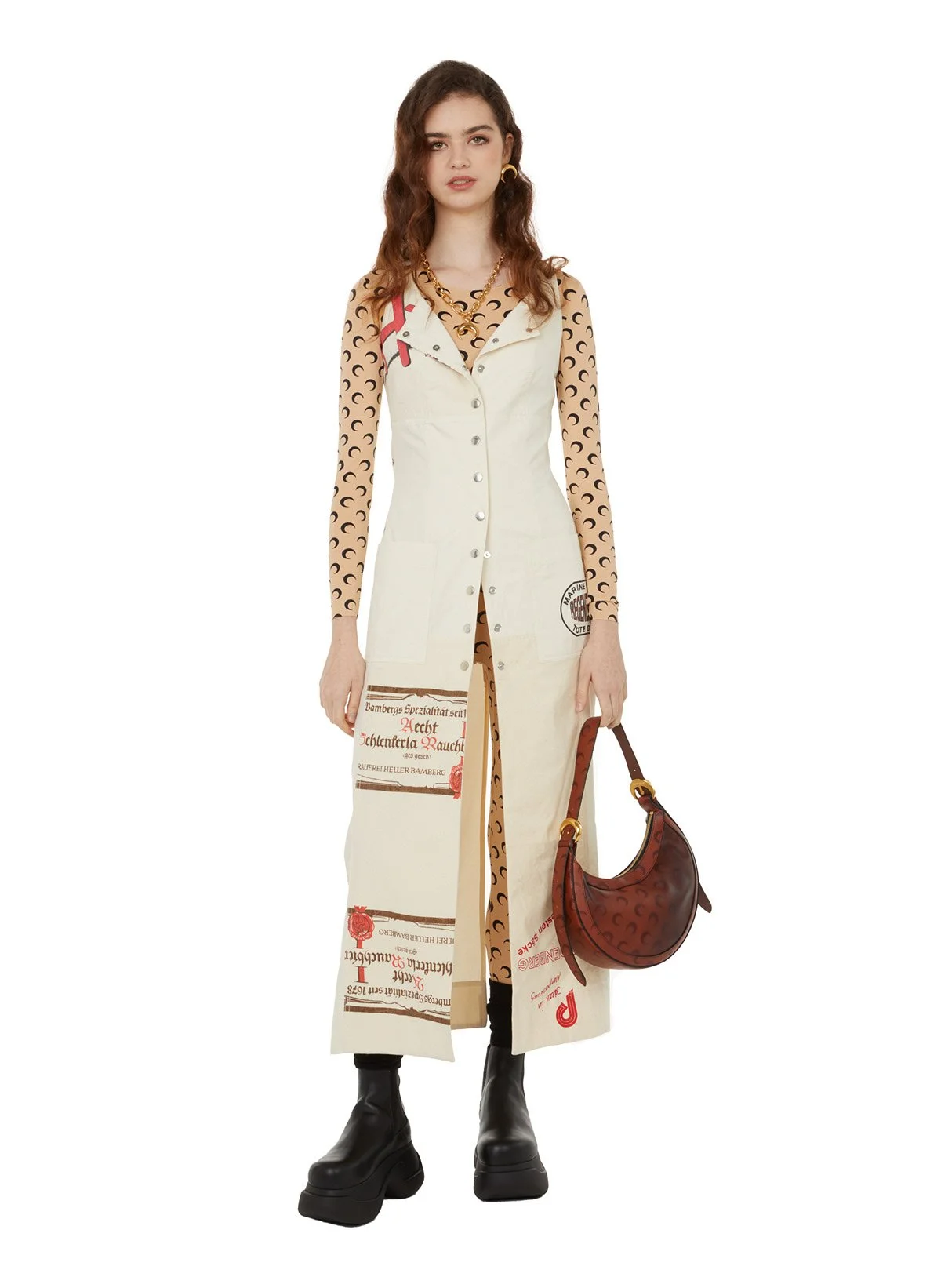 Une femme portant une robe longue avec des motifs de texte et d'images, un manteau sans manches beige, des bottes noires épaisses, des bijoux en or, et tenant un sac à main marron avec un motif
