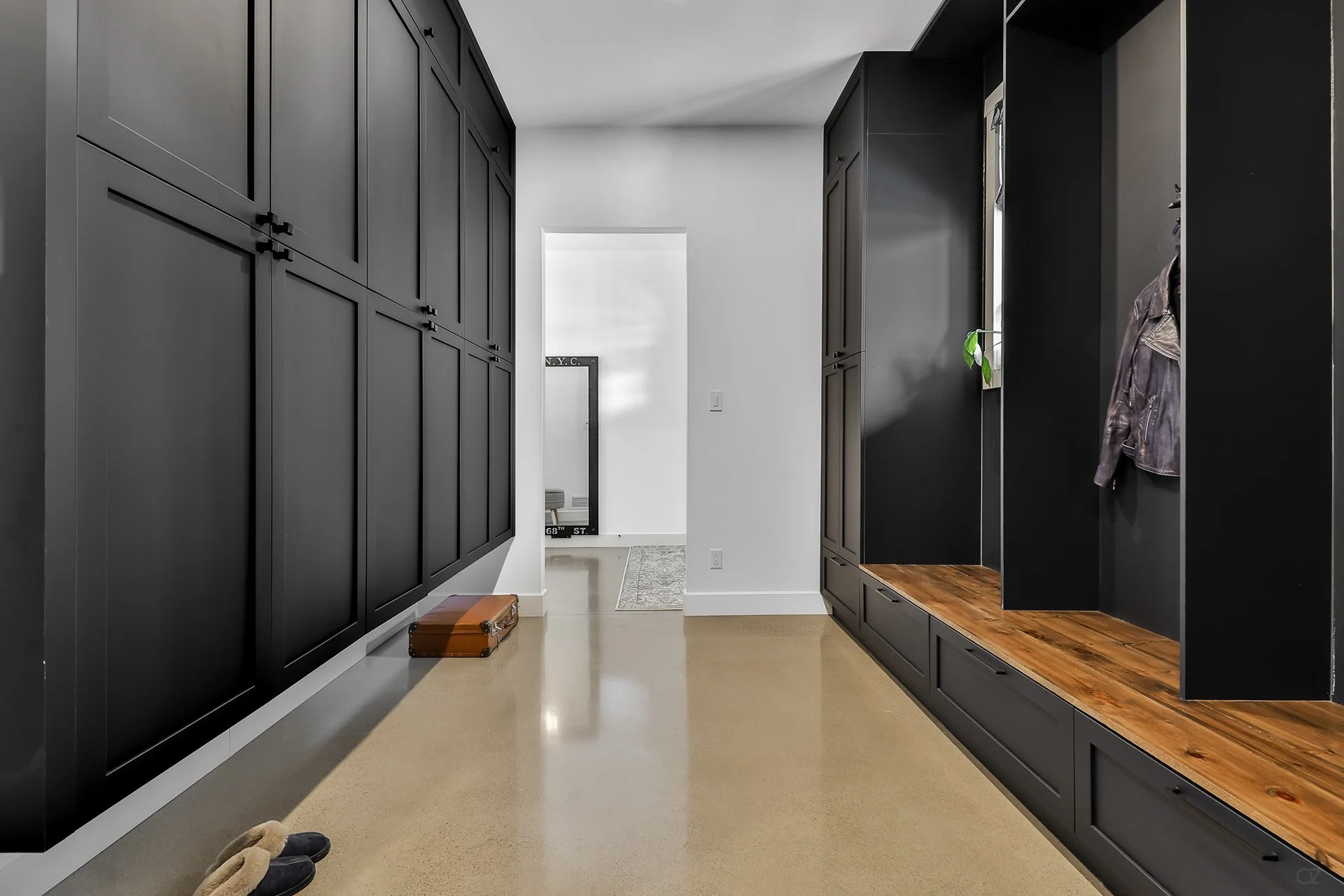 Entrée moderne avec armoires noires sur les murs, un sol beige, un miroir en arrière-plan, une porte ouverte menant à une salle d'entrée avec tapis, paire de chaussures à l'avant, une petite plante sur une galerie de l'entrée, une veste en cuir suspe