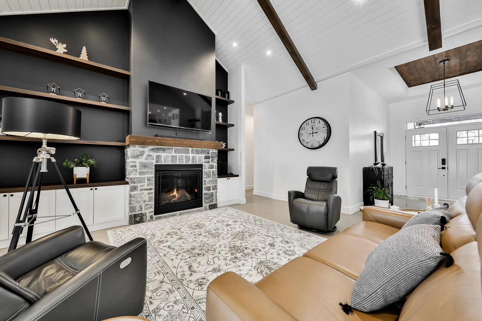 Salon moderne avec cheminée en pierre, fauteuils en cuir, tapis clair, étagères noires avec décorations, horloge ronde, pièce lumineuse avec grande porte d'entrée.