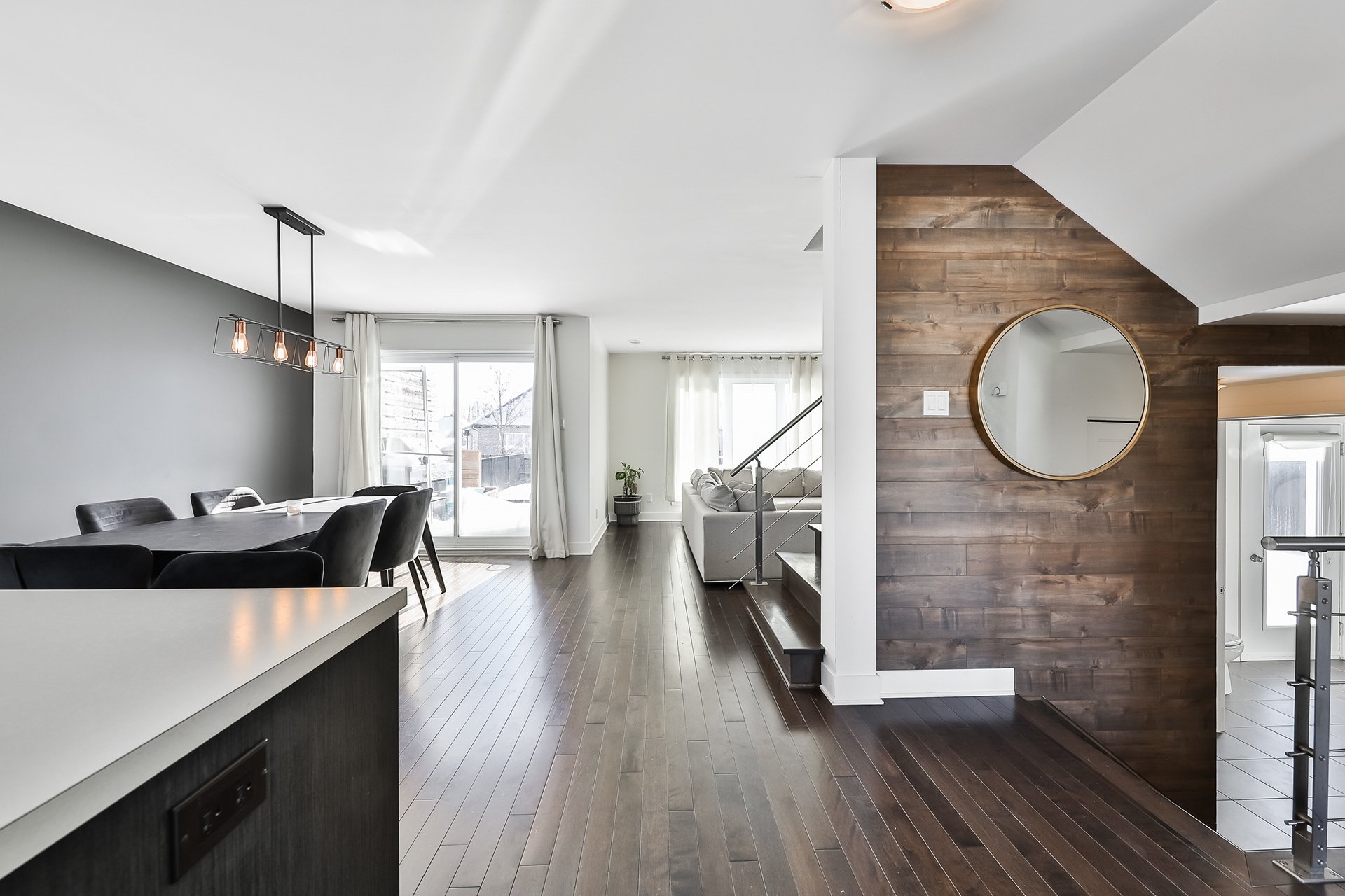 Intérieur d'une maison moderne avec salon et salle à manger, sol en bois foncé, mur en bois clair, grande fenêtre avec rideaux blancs, miroir rond sur le mur en bois, éclairage suspendu au plafond