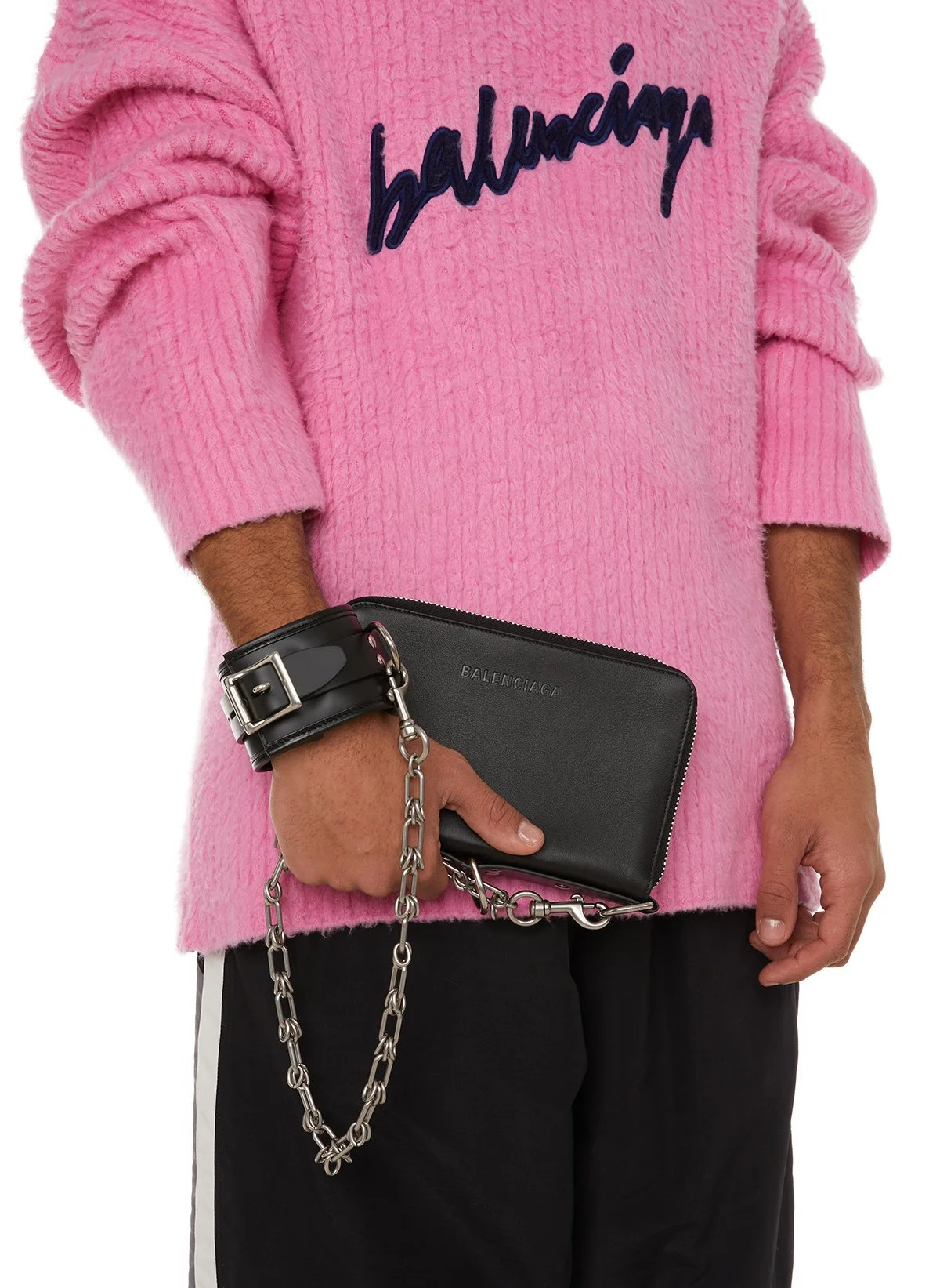 Personne portant un pull rose avec l'inscription "balenciaga", tenant un portefeuille noir avec une chaîne en métal, et portant un bracelet noir.