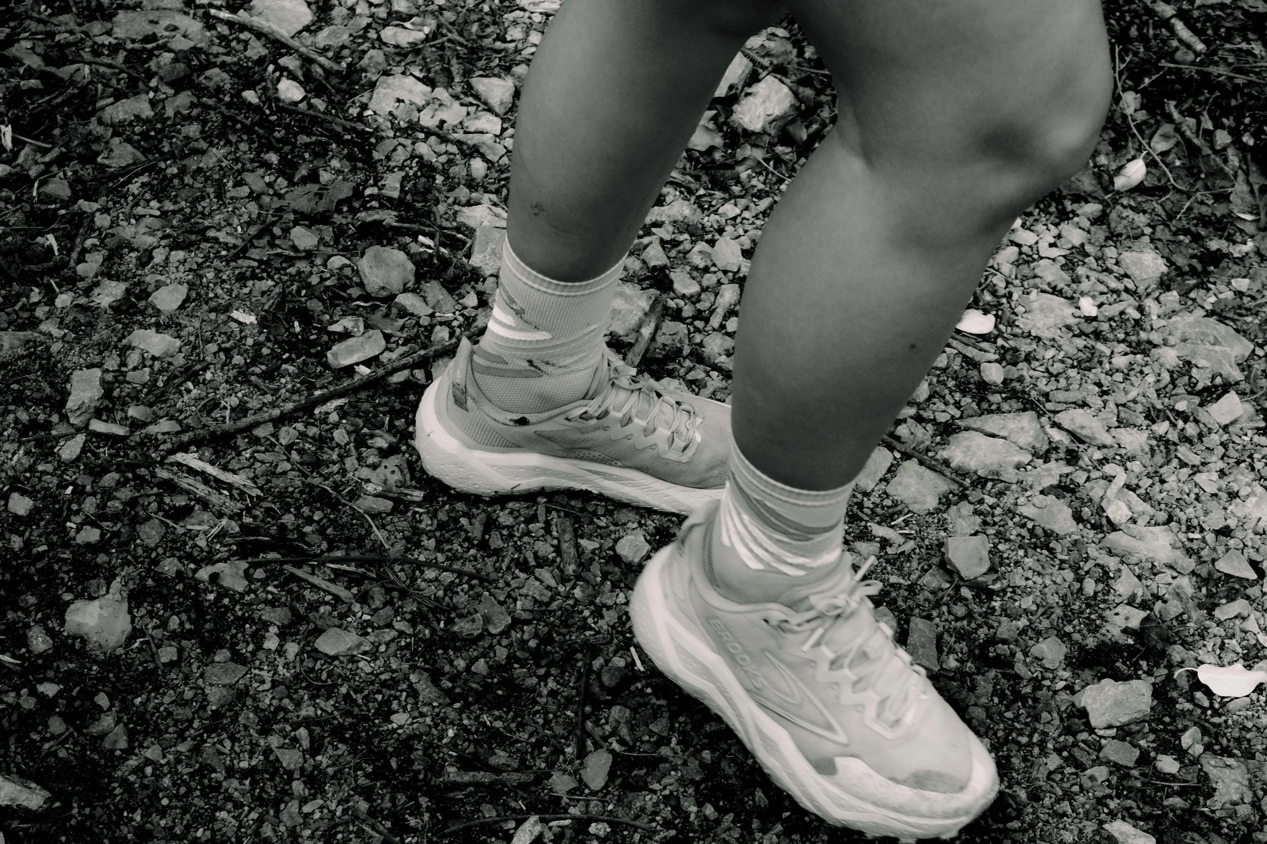 Jambes et chaussures d'une personne debout sur un sol rocheux, en noir et blanc.
