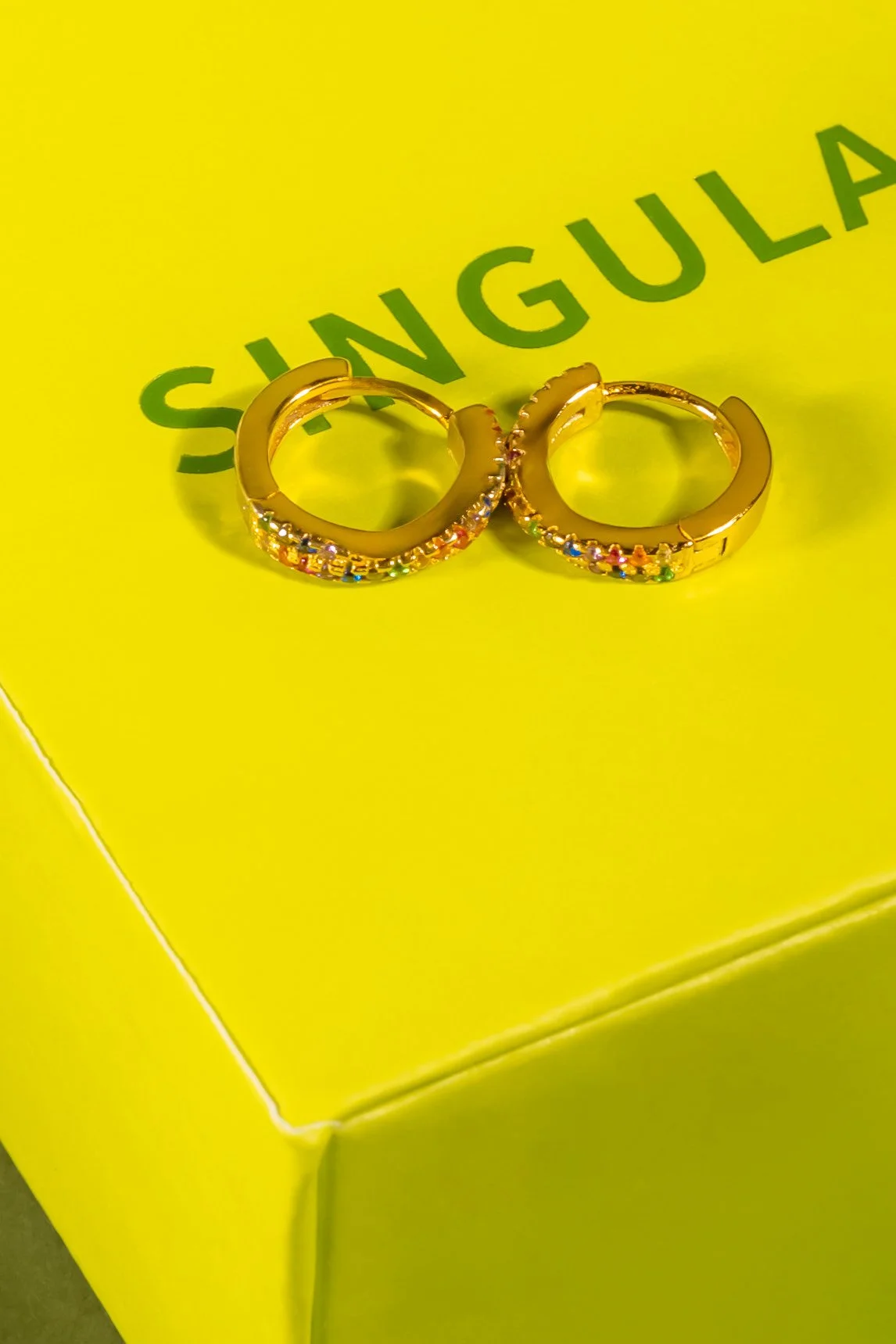 Une paire de petits anneaux en or avec des pierres scintillantes colorées, posée sur un fond jaune avec le mot 'SINGULA' écrit en vert.