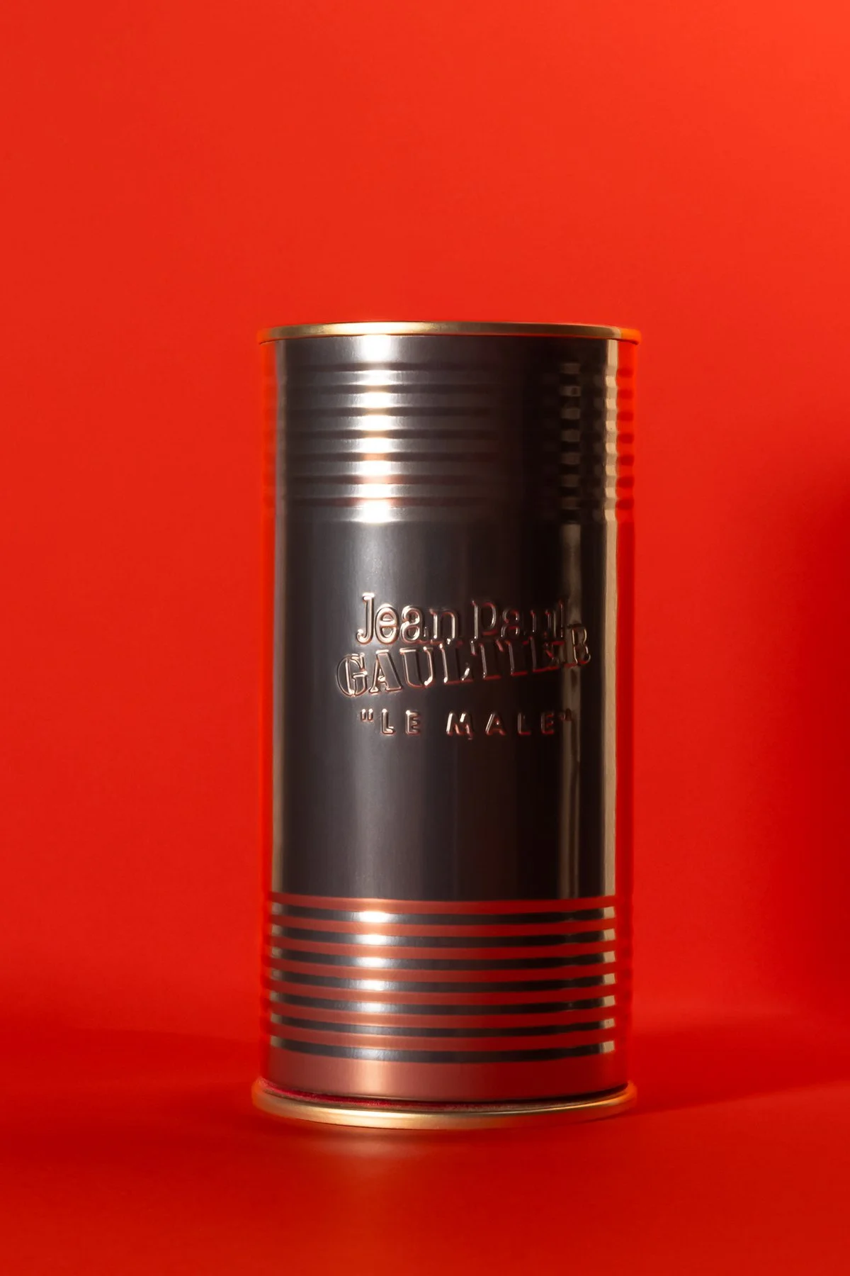 Une boîte en métal noire avec des bandes horizontales et l'inscription "Jean Paul Gaultier Le Male" en relief, posé sur un fond rouge