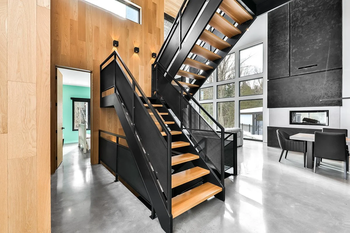 Intérieur moderne d'une maison avec escalier en métal noir, marches en bois clair, murs en bois et noir, grande baie vitrée laissant entrer la lumière, cheminée encastrée, coin repas avec table et chaises noires.