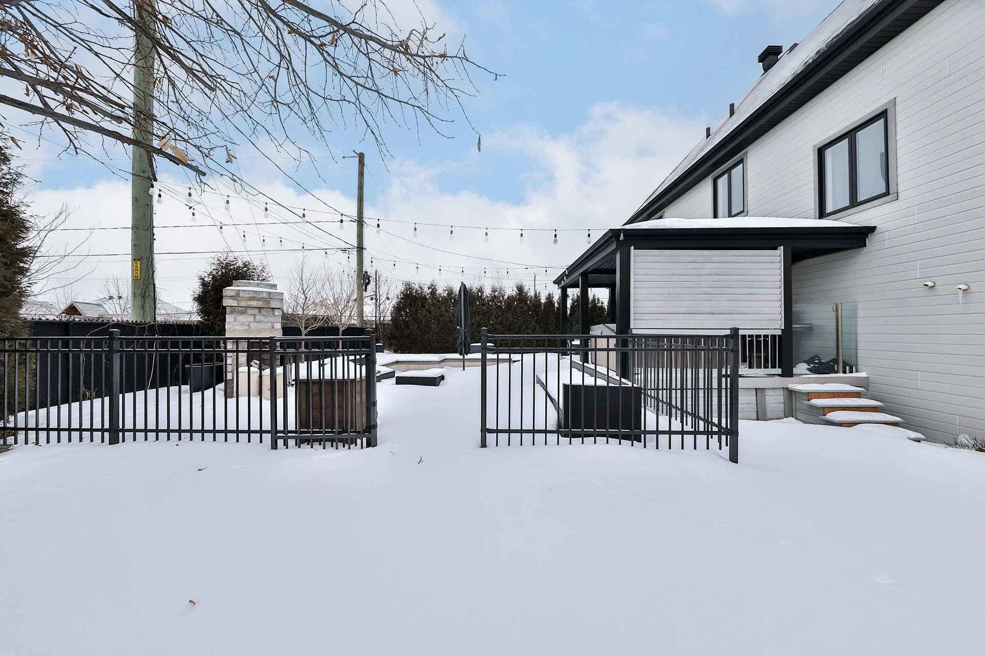 Une maison moderne sous la neige avec une clôture en métal noir, des escaliers en bois recouverts de neige, une terrasse et un ciel bleu avec quelques nuages.