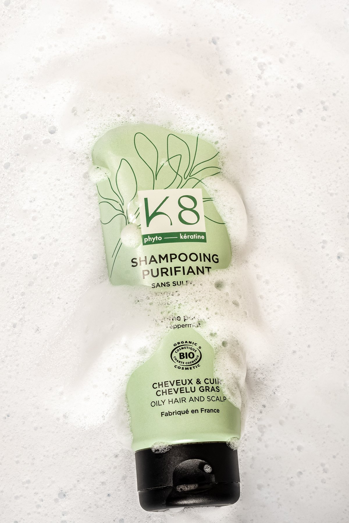Un tube de shampooing purifiant pour cheveux gras et cuirs chevelus gras, de couleur vert clair, avec un bouchon noir, flottant dans de l'eau mousseuse.