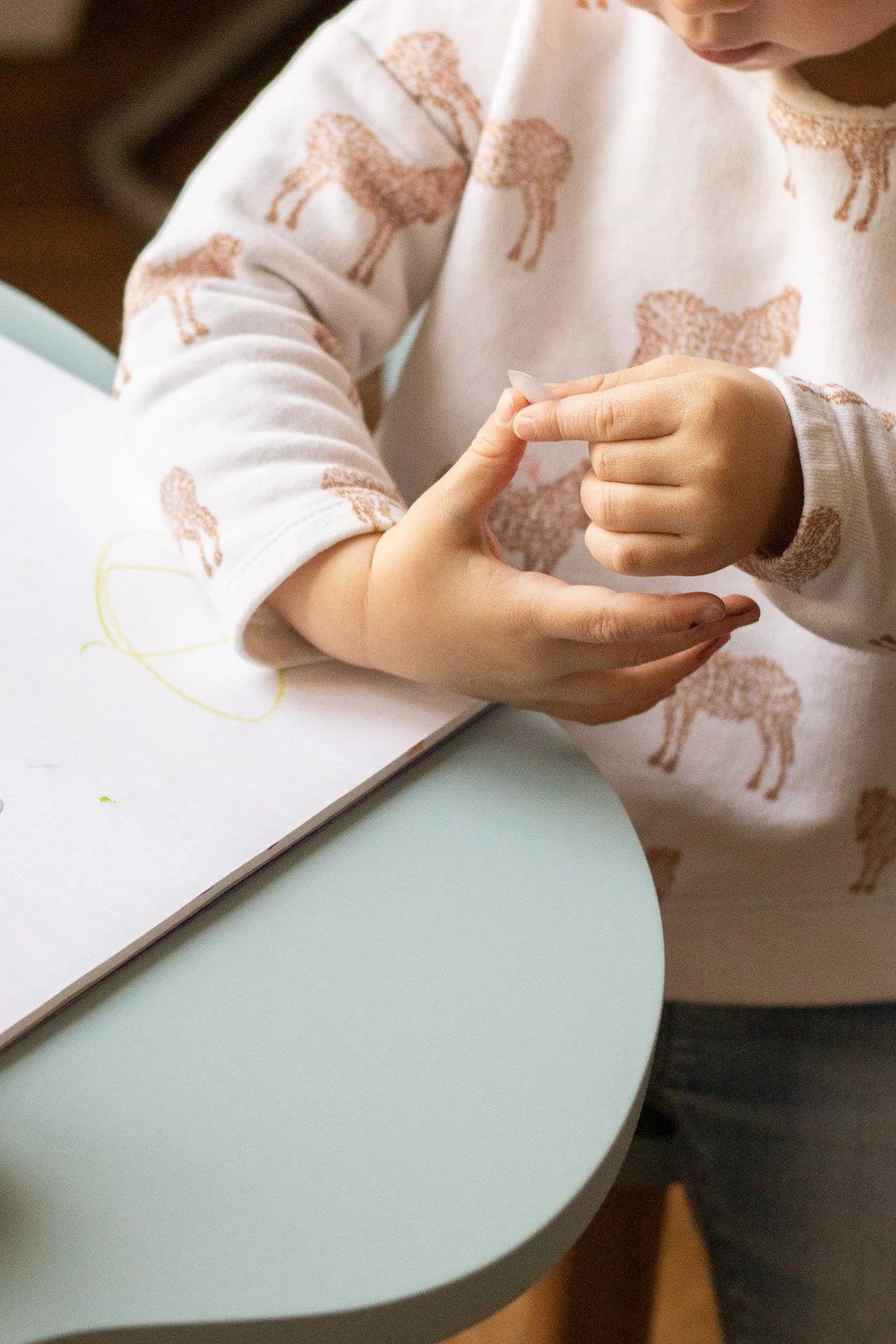 Un enfant en pull à motif girafes, faisant un geste avec ses mains au-dessus d'une table blanche avec un cahier ouvert. La scène semble se dérouler à l'intérieur.