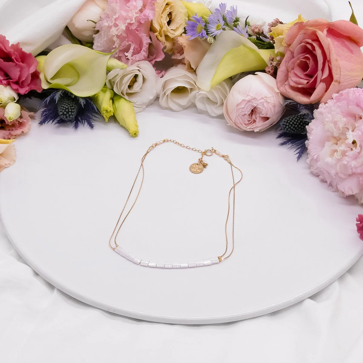 Collier delicat avec pendentif en or, posé sur une assiette blanche, entouré de fleurs roses, blanches, et violettes.