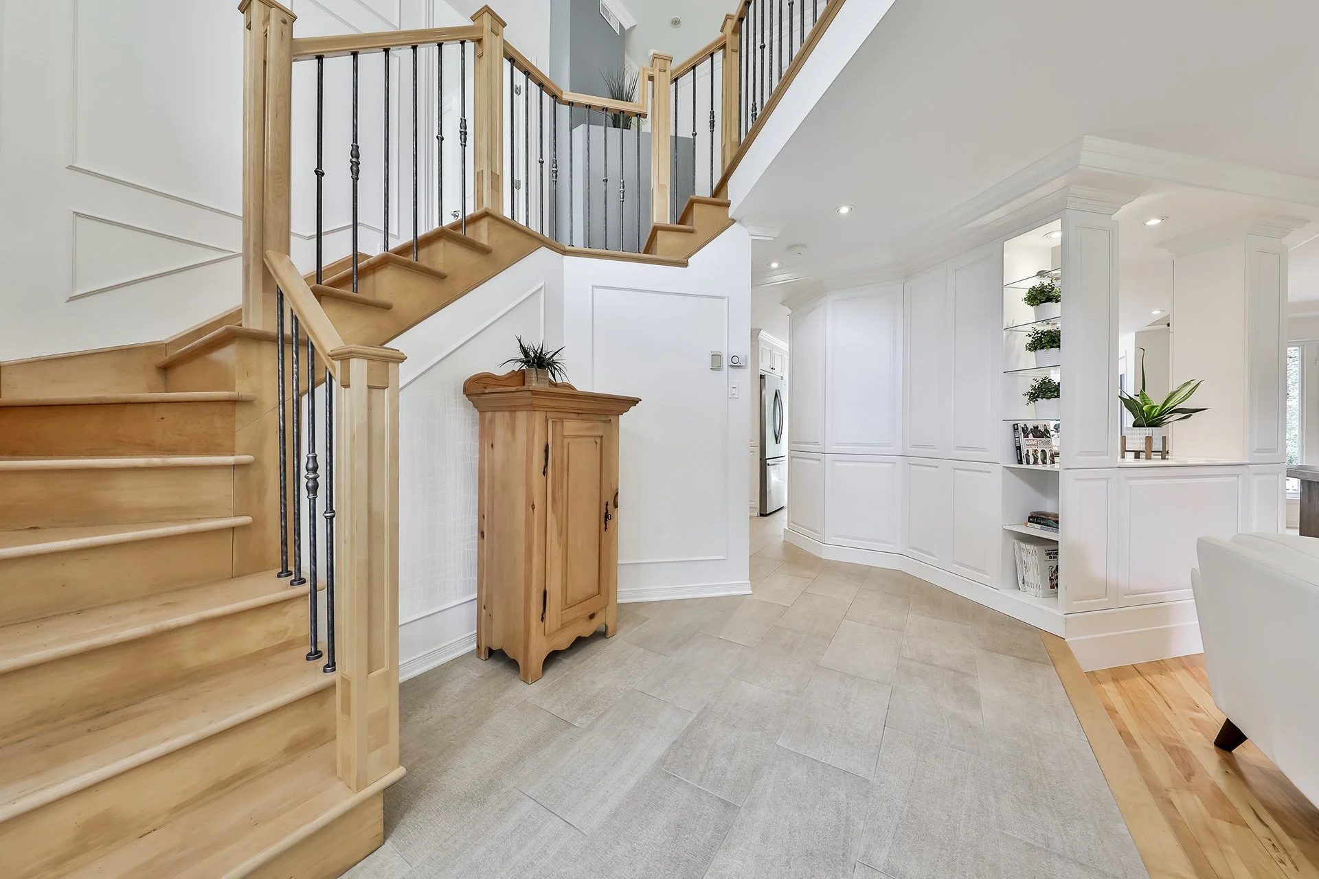 Intérieur d'une maison avec escalier en bois clair, mur blanc, armoires encastrées, et étagères avec des plantes et des livres.
