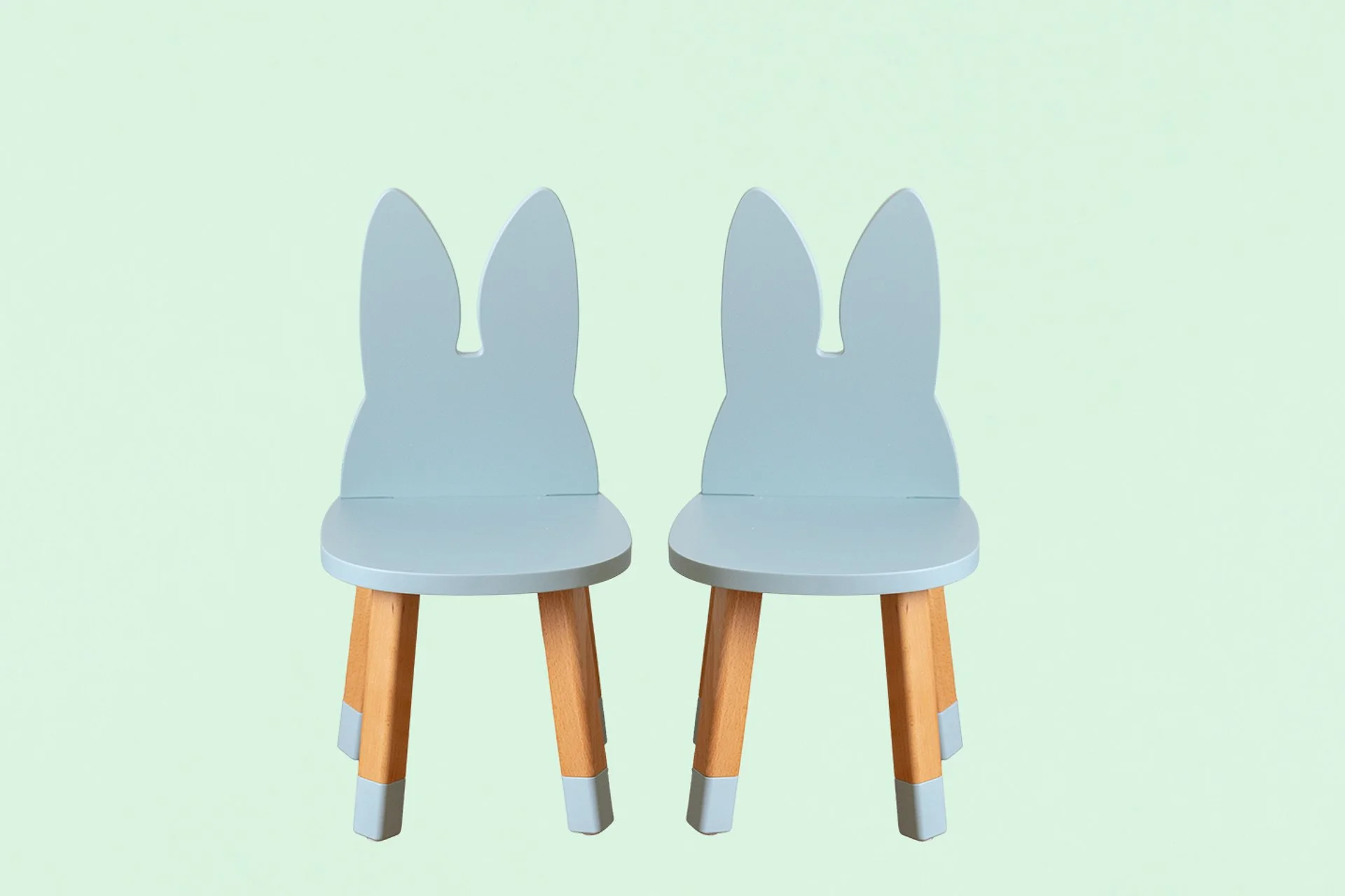 Deux chaises pour enfants avec dossiers en forme de lapin, en bois avec assises grises, contre un fond vert pastel.