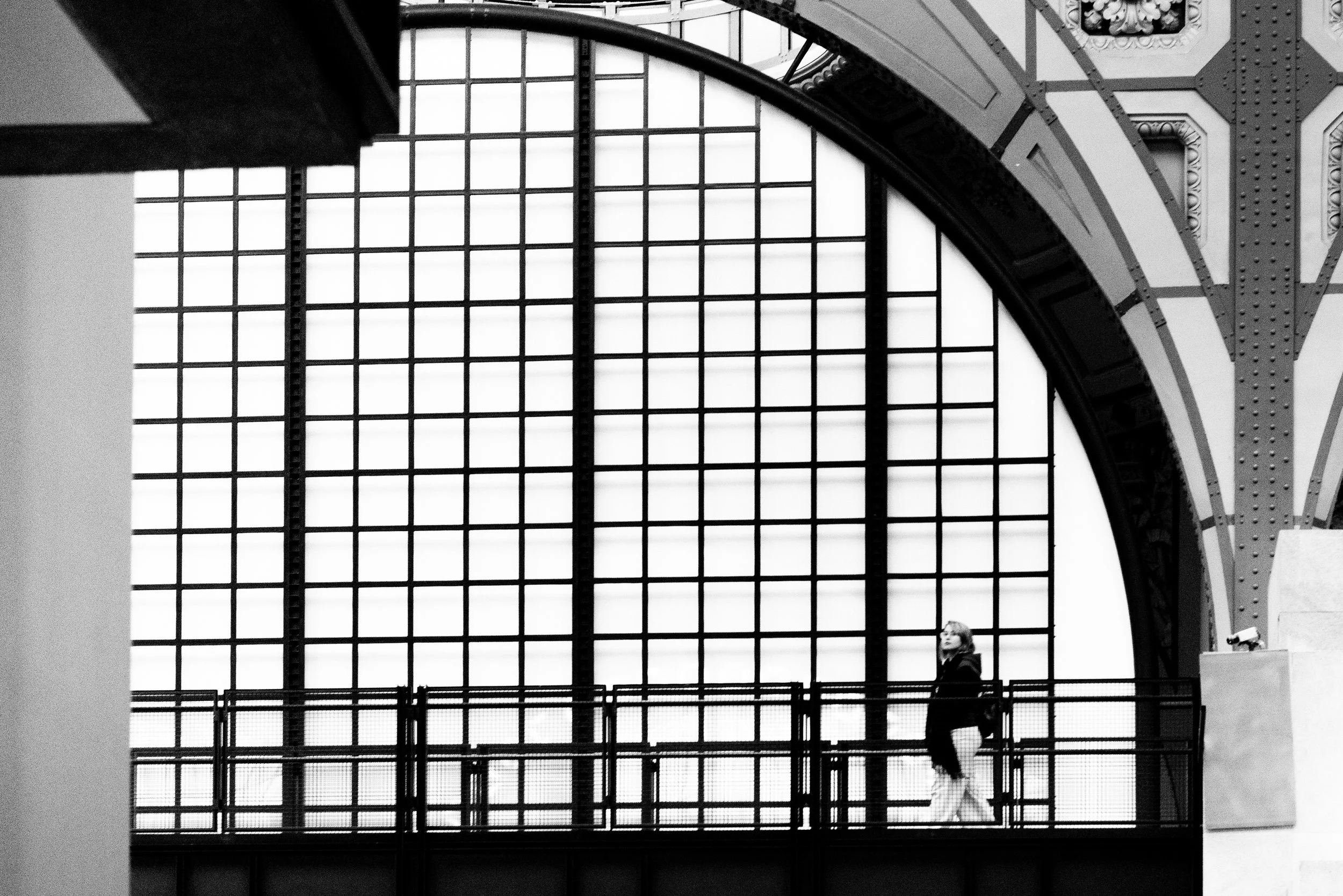 Une femme marche dans une galerie industrielle avec un plafond en arc et une grande fenêtre en grille en arrière-plan, en noir et blanc.
