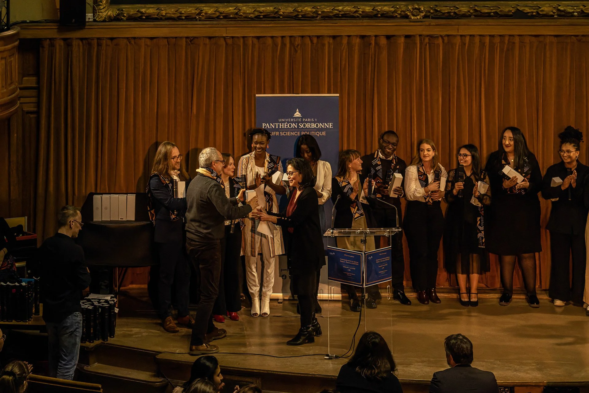 Cérémonie de remise de diplômes ou d'une distinction à l'université, avec un groupe de personnes sur scène, lauréats et organisateurs, dans une salle avec rideaux en bois, une bannière indiquant 'Université Paris 1 Pantheon Sorbonne, Sciences Politiq