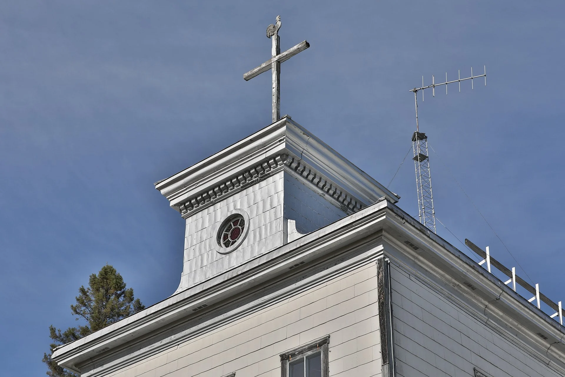 Vue de la partie supérieure d'une ancienne église en bois blanc avec une croix en bois sur le toit et une petite fenêtre ronde à vitre colorée, ciel bleu en arrière-plan.