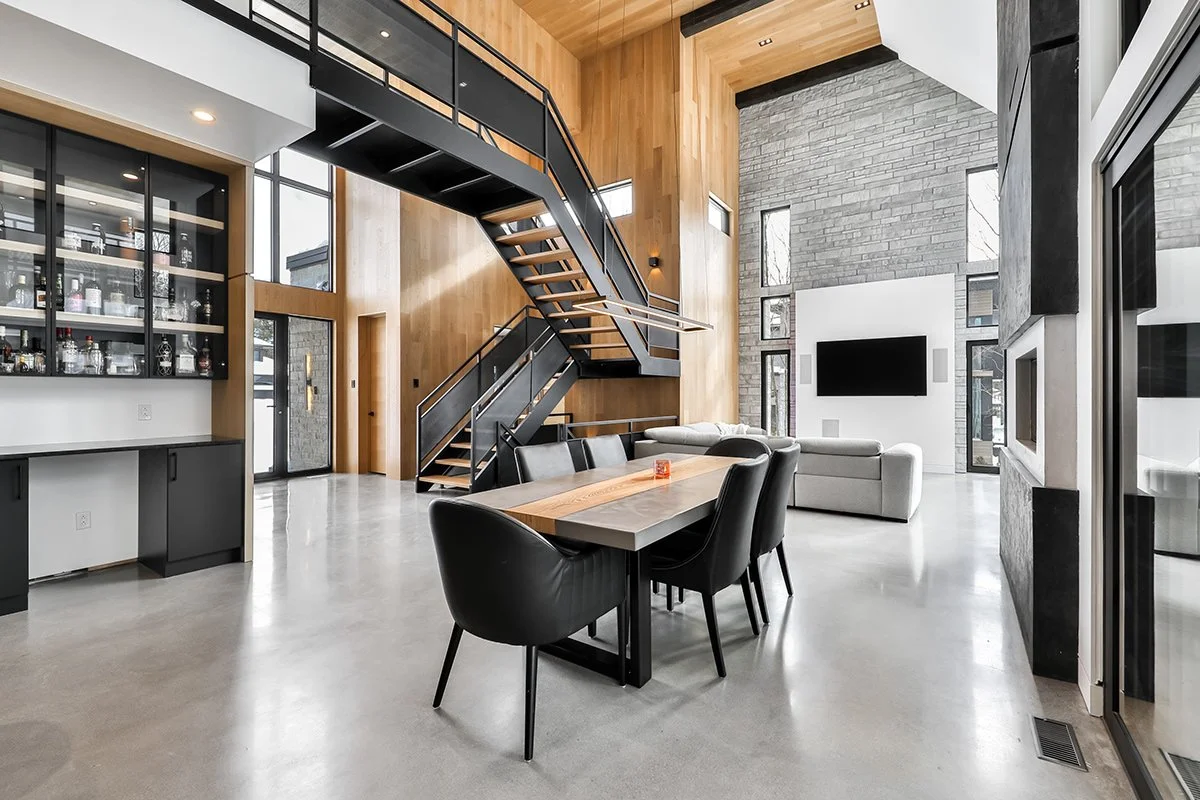 Un salon moderne avec un mobilier élégant, un mur en pierre avec une grande télévision, un escalier en métal et bois, et de grandes fenêtres laissant entrer la lumière naturelle.