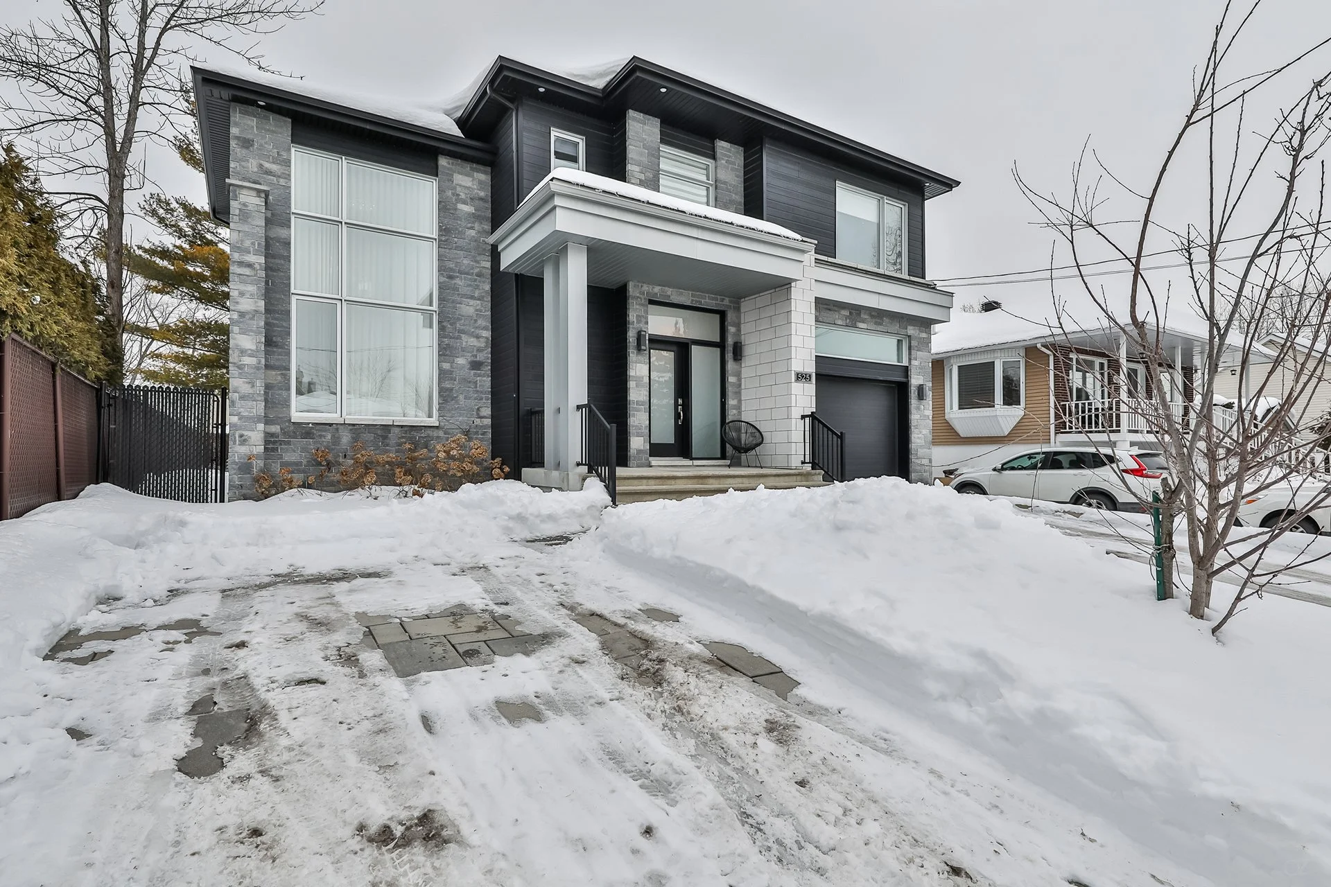 Maison moderne à plusieurs étages en hiver, entourée de neige, avec une allée enneigée et quelques arbres nus.