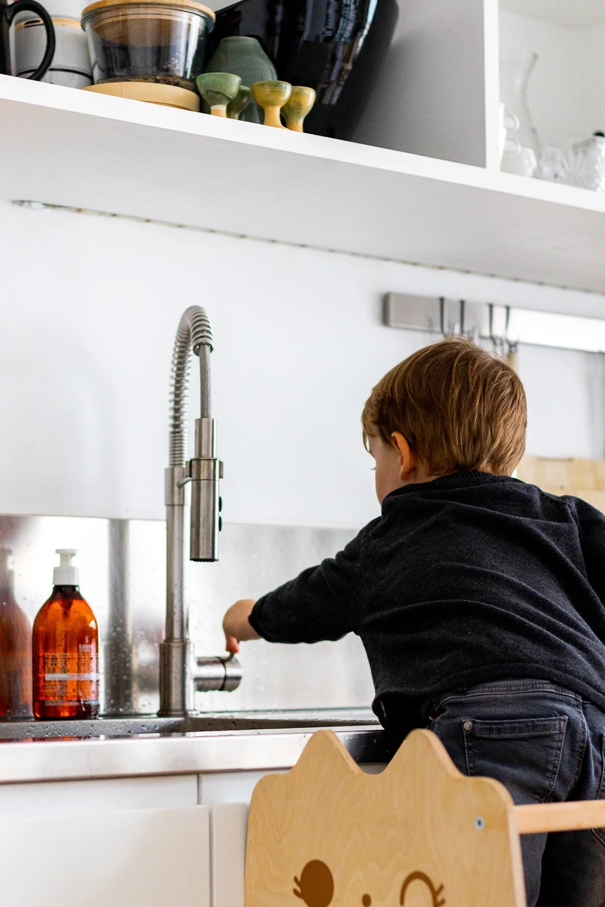 Un enfant avec des cheveux bruns, vu de dos, joue dans une cuisine moderne près de l'évier, avec une bouteille de savon orange à gauche.