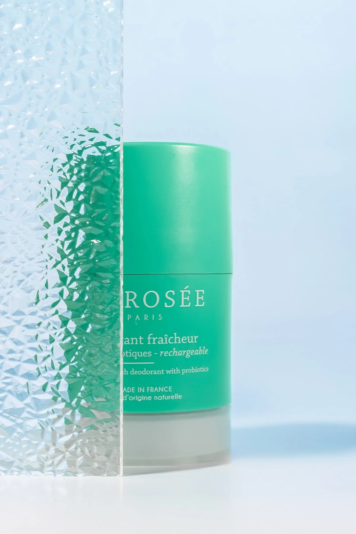Un déodorant en stick vert de la marque Rosée Paris, mettant en avant sa formule fraîche et rechargeable, avec un fond clair et un objet en verre texturé en premier plan.