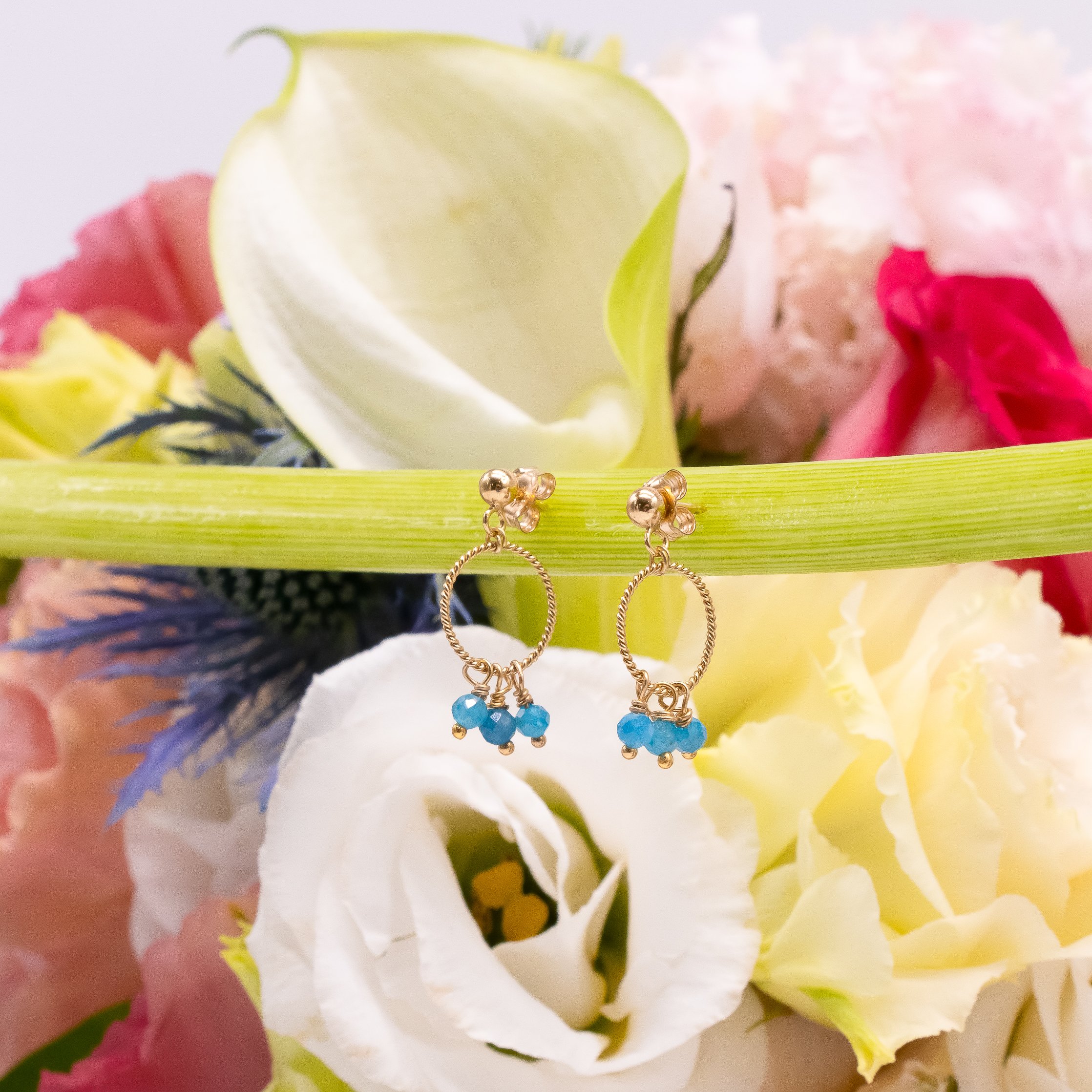 Boucles d'oreilles en or avec petites perles bleues suspendues, placées sur une tige verte, avec un bouquet de fleurs colorées en arrière-plan.