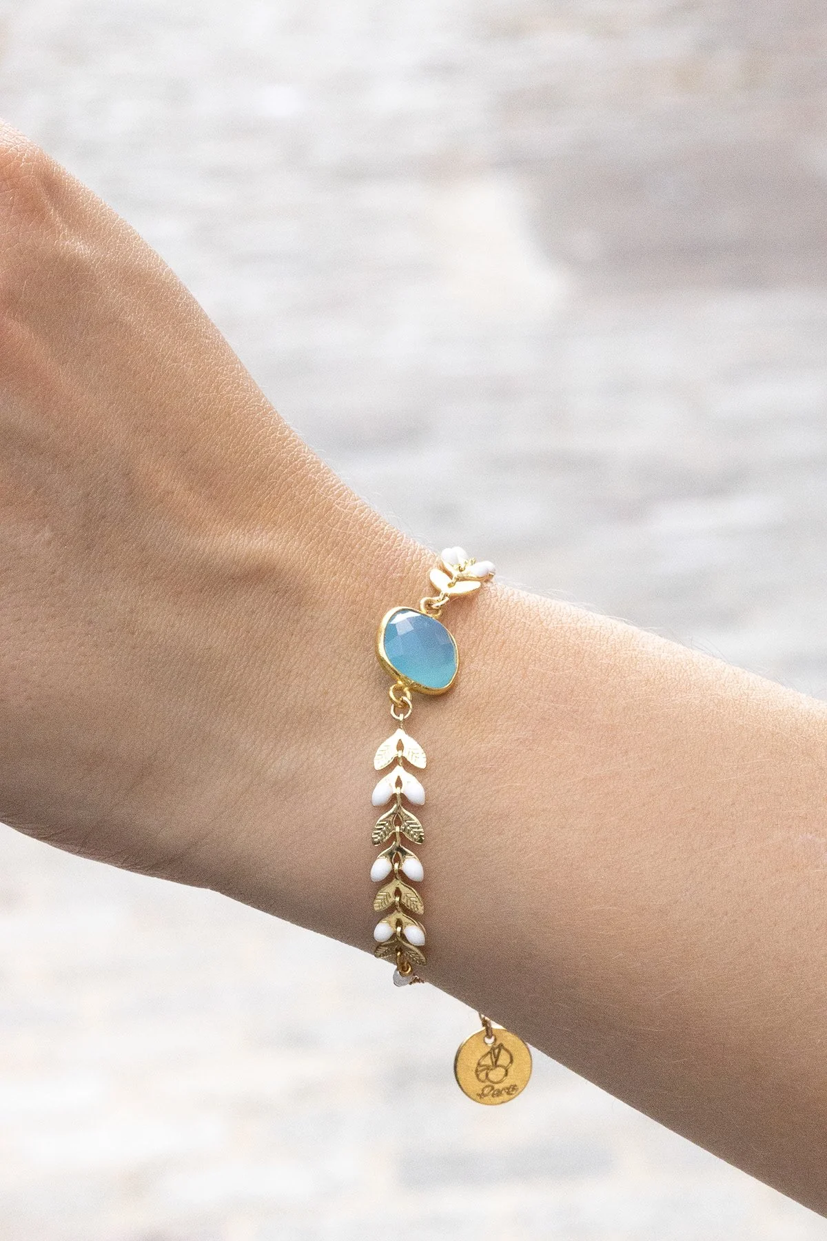 Bracelet doré avec un pendentif bleu en pierre et des charms en forme de feuilles blanches et dorées, avec un petit pendentif rond gravé de lettres mystérieuses.