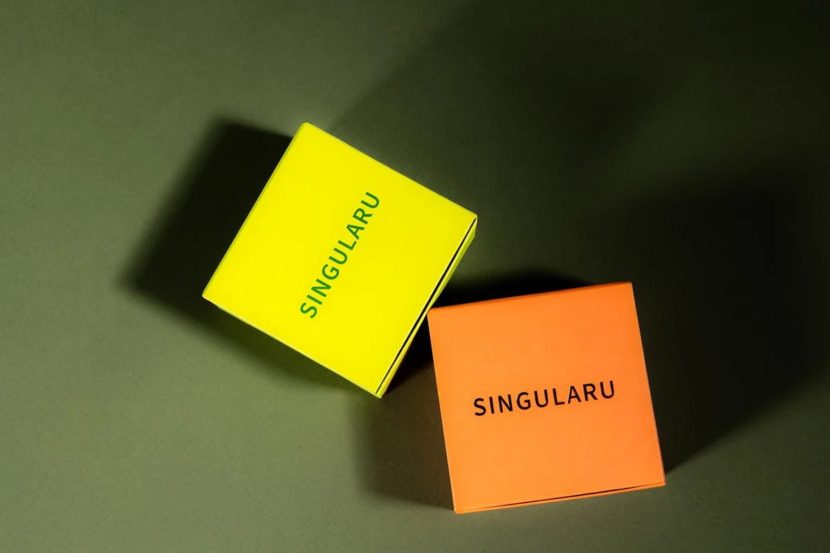 Deux boîtes colorées portant le mot "SINGULARU" sur un fond gris. La boîte à gauche est jaune, celle à droite est orange.