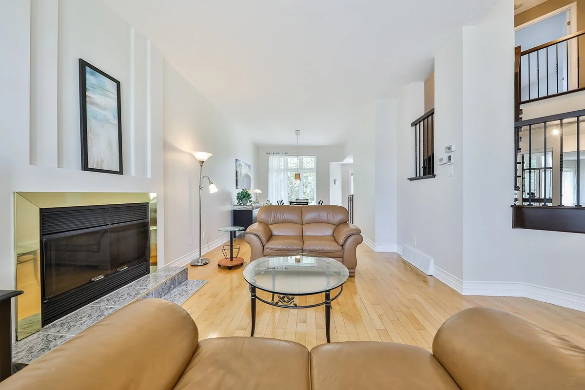Salon lumineux avec canapé en cuir beige, cheminée à cadre or, mobilier en bois clair, mur blanc et parquet en bois, vue sur salle à manger avec fenêtre.