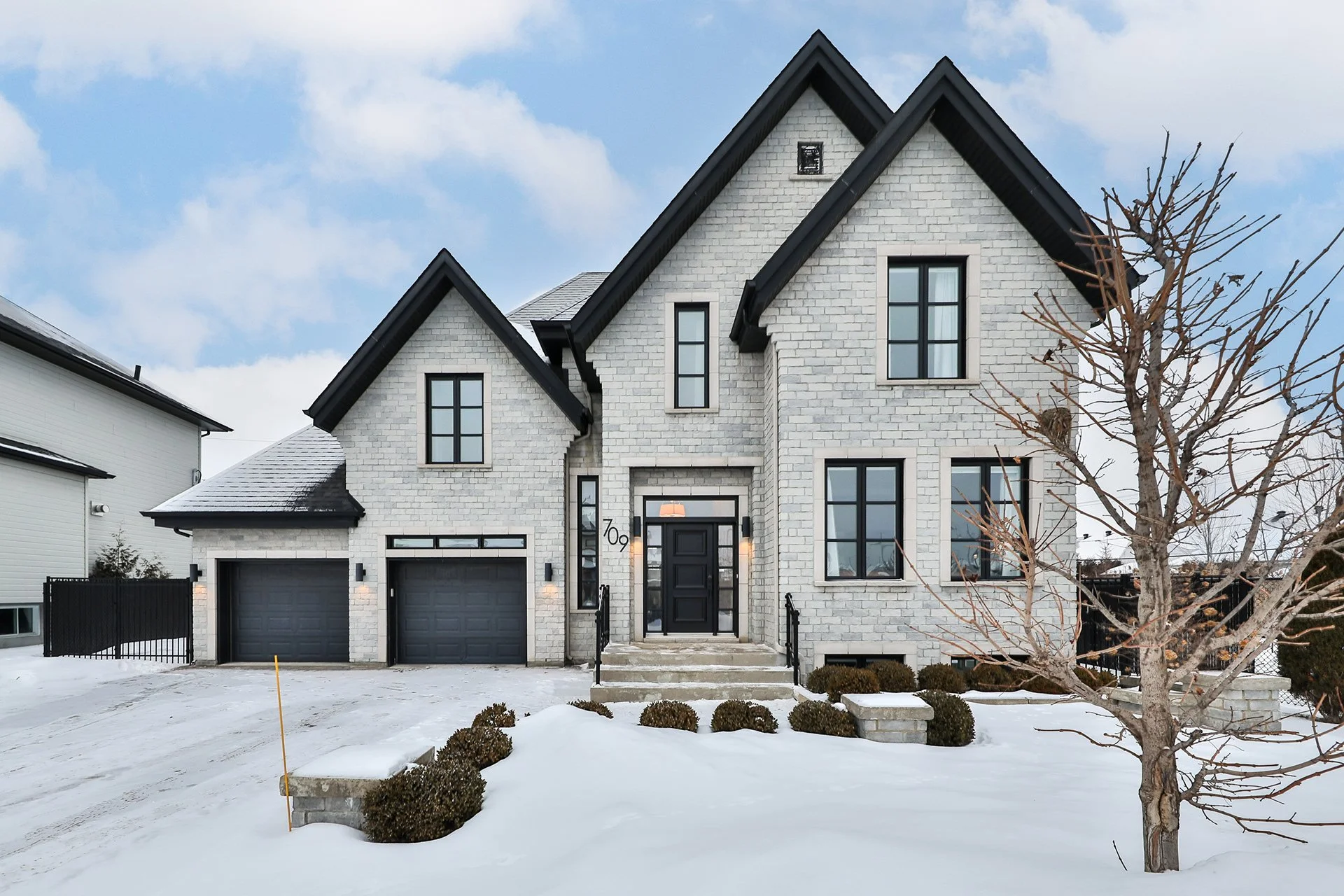 Maison moderne à deux étages avec façade en briques blanches, toiture noire, garage double, arbres dénudés, et neige au sol, sous un ciel partiellement nuageux.