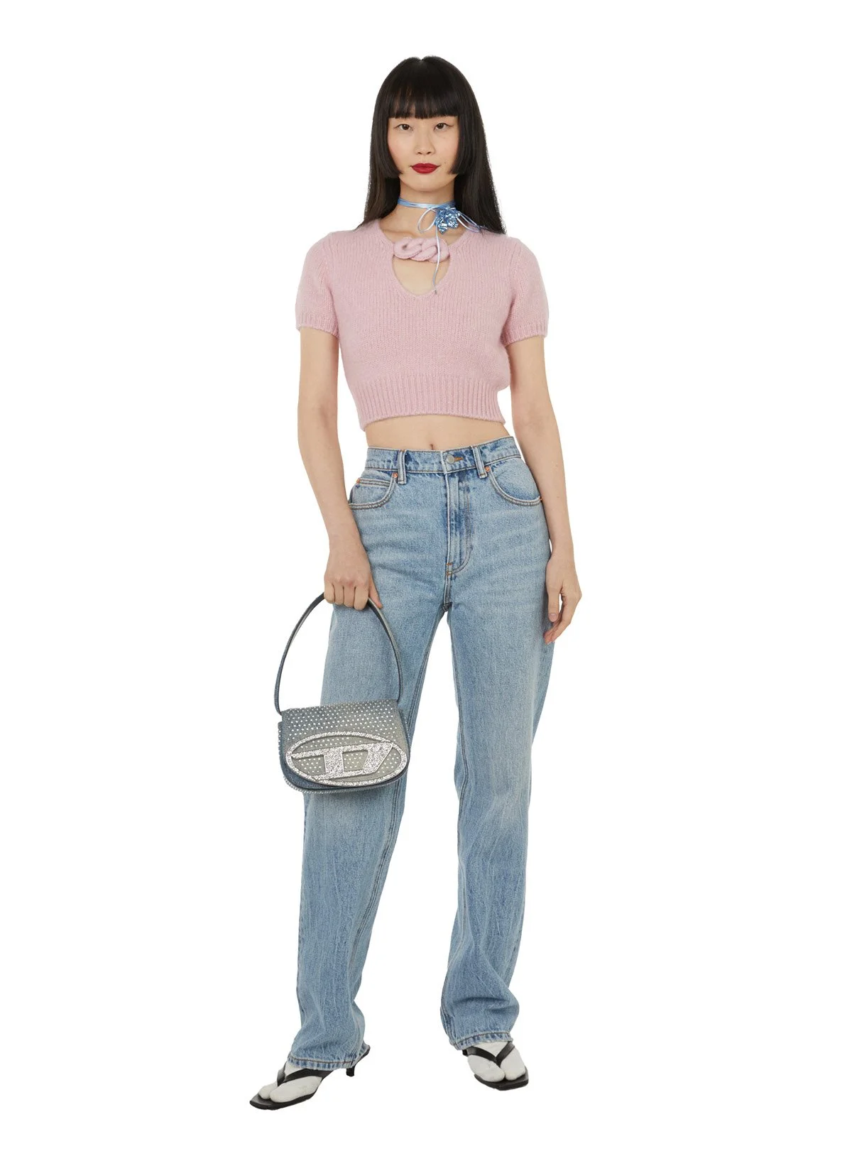 Jeune femme portant un haut en tricot rose, un jean décontracté, des chaussures noires et blanches, tenant un sac à main gris avec des détails en strass, avec un fond blanc.