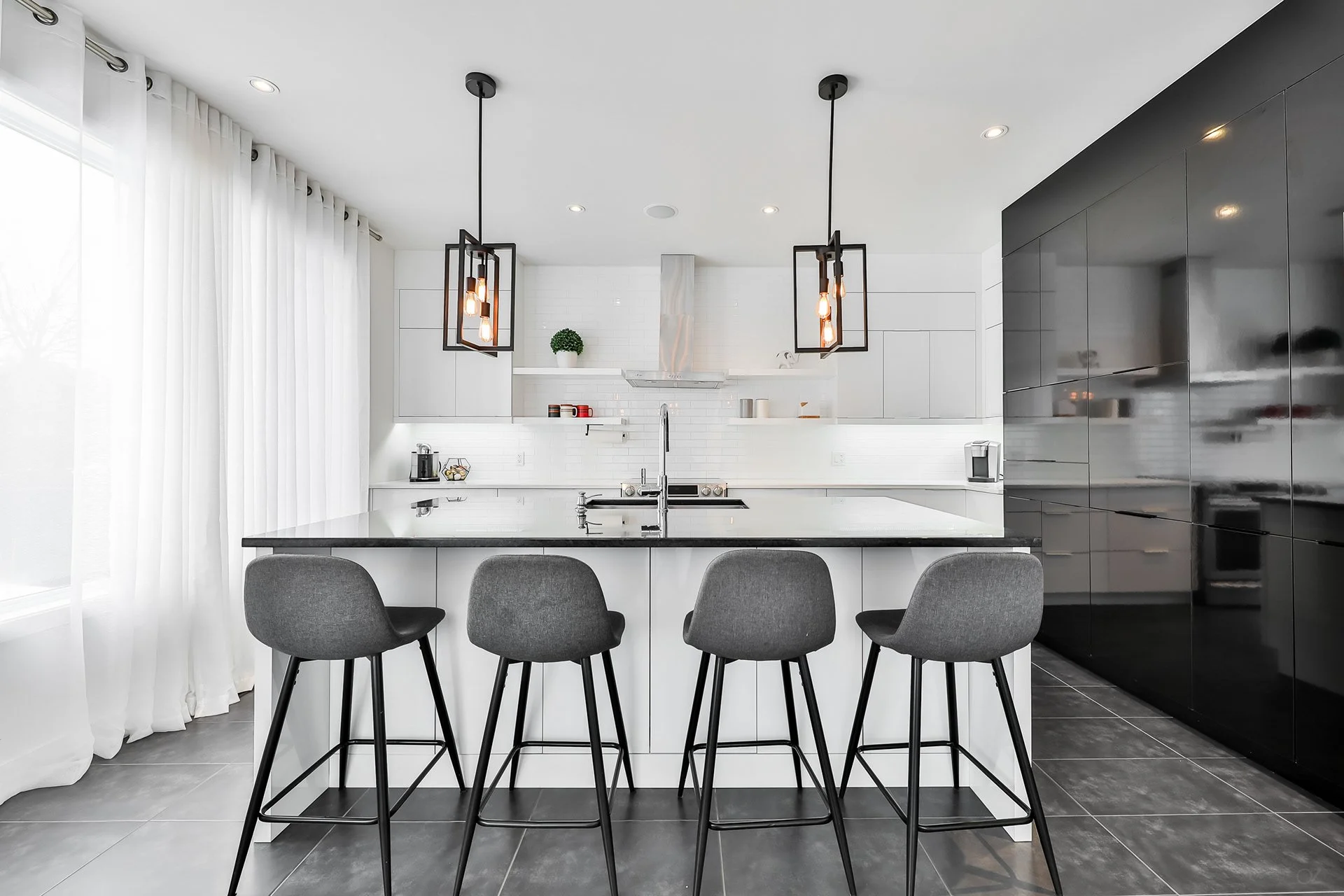 Cuisine moderne noire et blanche avec îlot central, quatre tabourets gris, pendant suspendu noir, portes de placard noires, et fenêtre avec rideaux blancs.