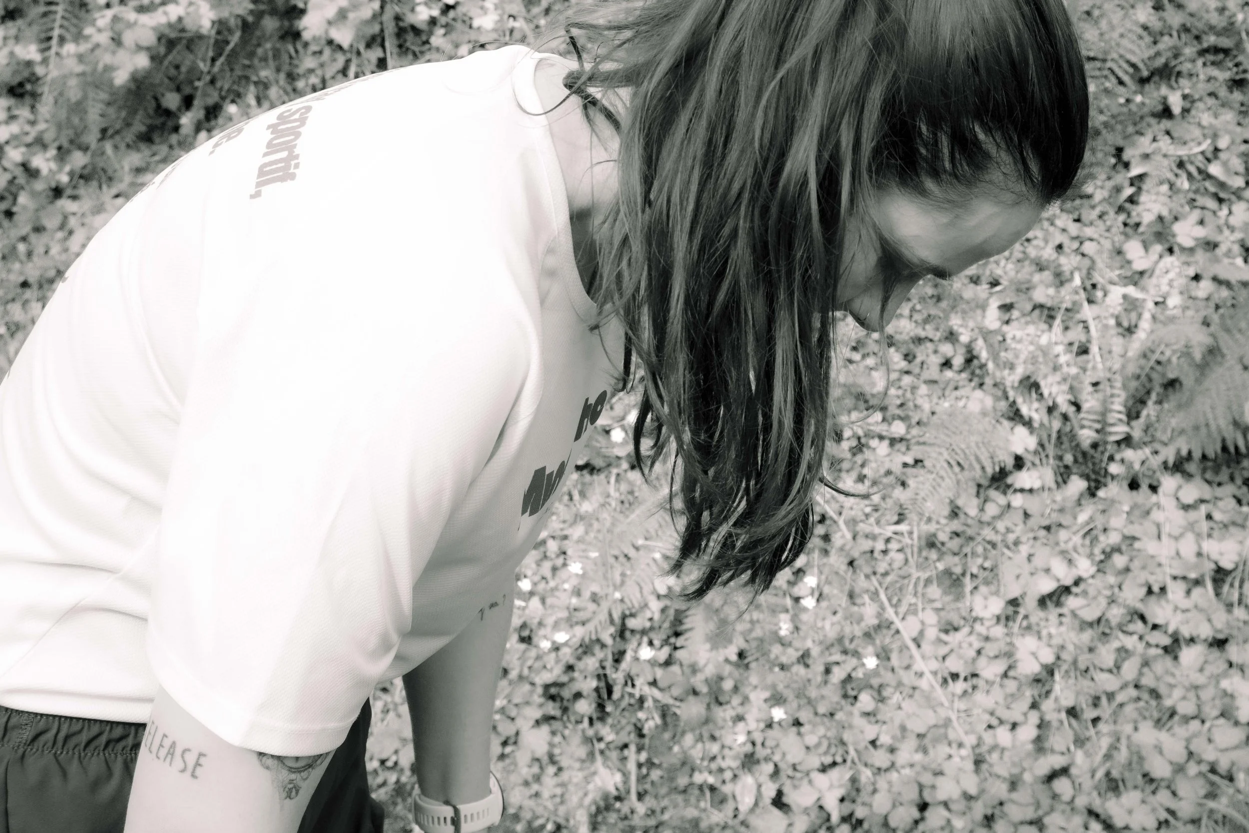 Une personne avec de longs cheveux bruns, portant un t-shirt blanc, inclinée en avant dans une zone boisée avec des feuilles et des fougères.