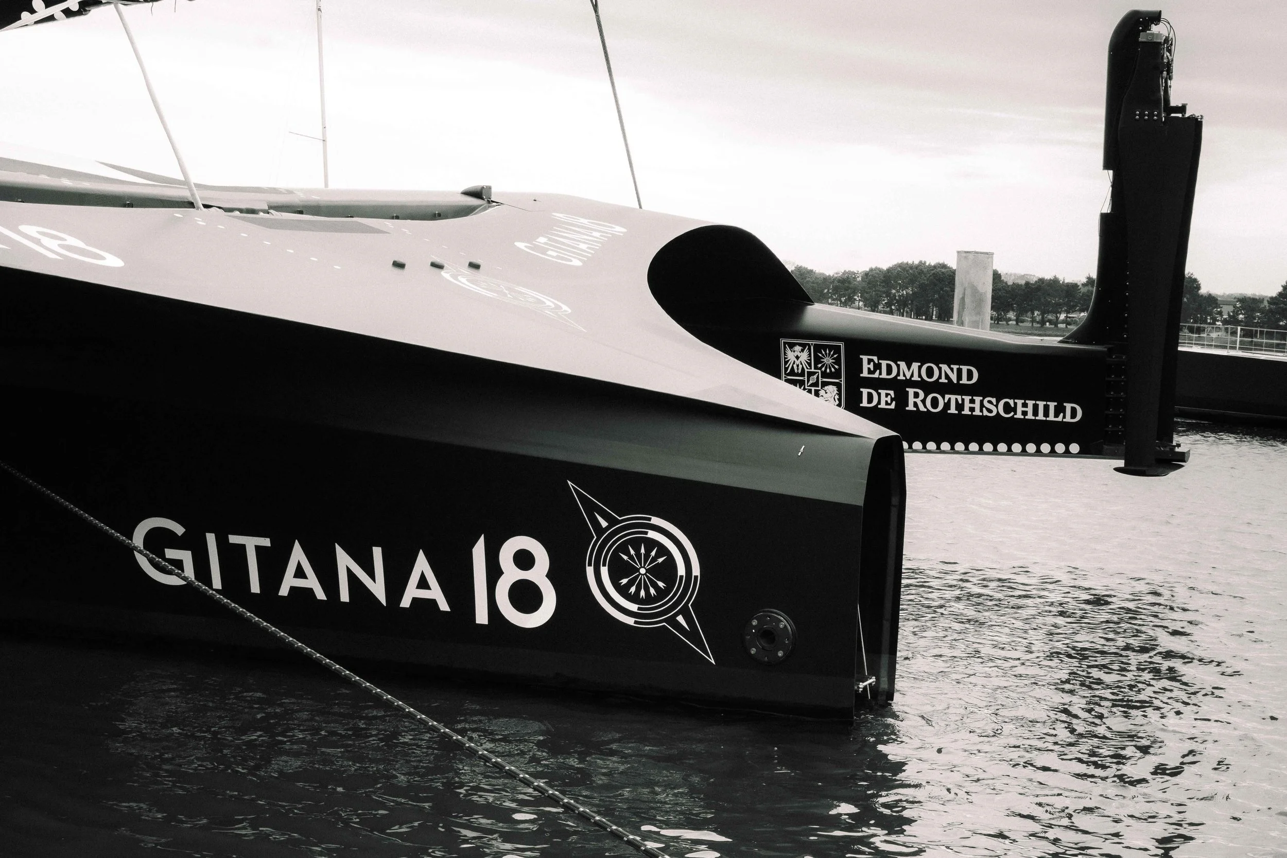 Deux voiliers de course amarrés dans un port, dont un portant le nom 'GITANA 18' et le numéro 18, avec des logos et des noms de sponsors, dont 'EDMOND DE ROTHSCHILD'.