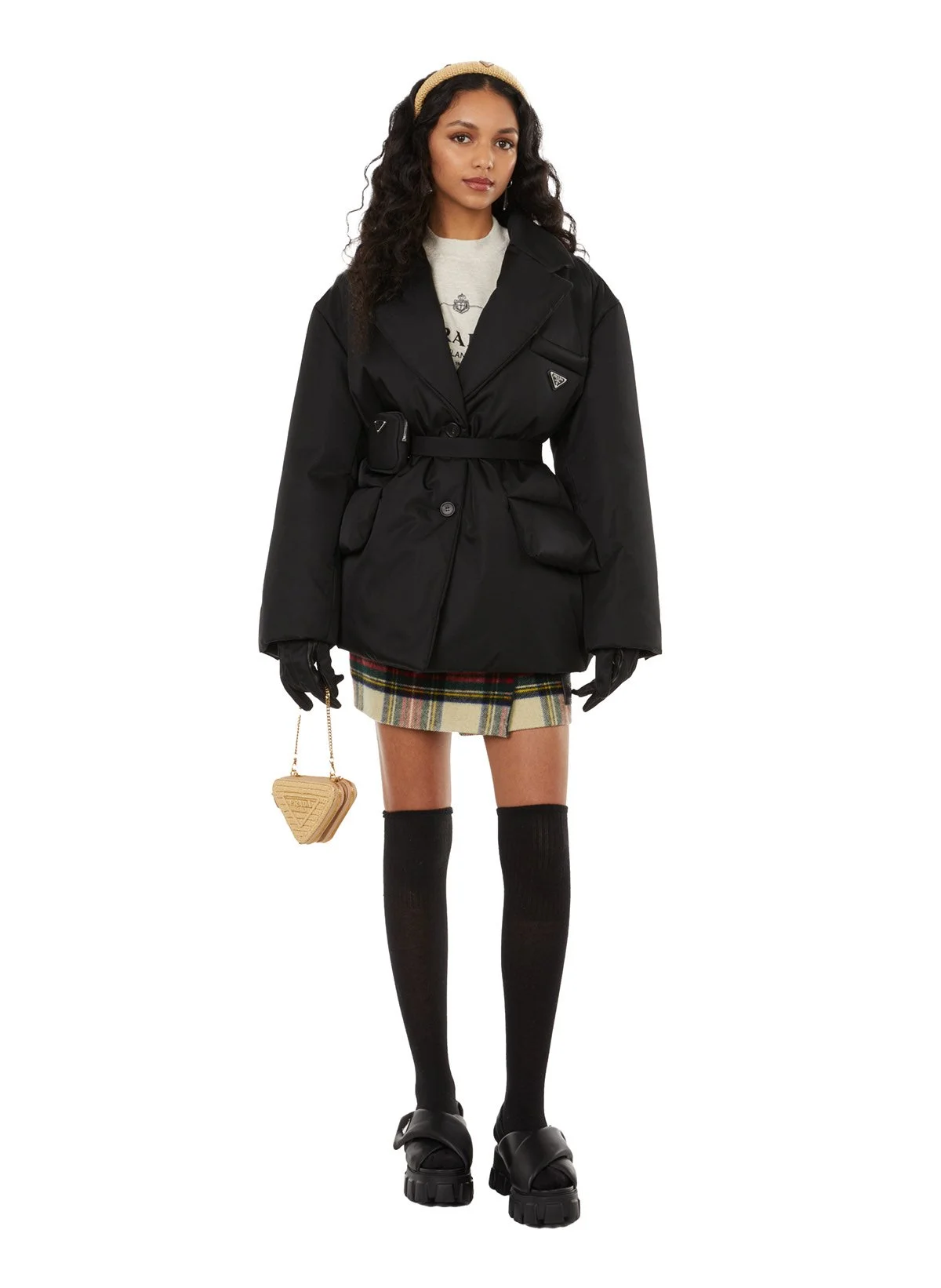 Une femme porte un manteau noir, une jupe tartan, des chaussettes hautes noires, des sandales épaisses, et tient un petit sac beige en forme de cœur. Elle a des cheveux bouclés et porte un bandeau beige.