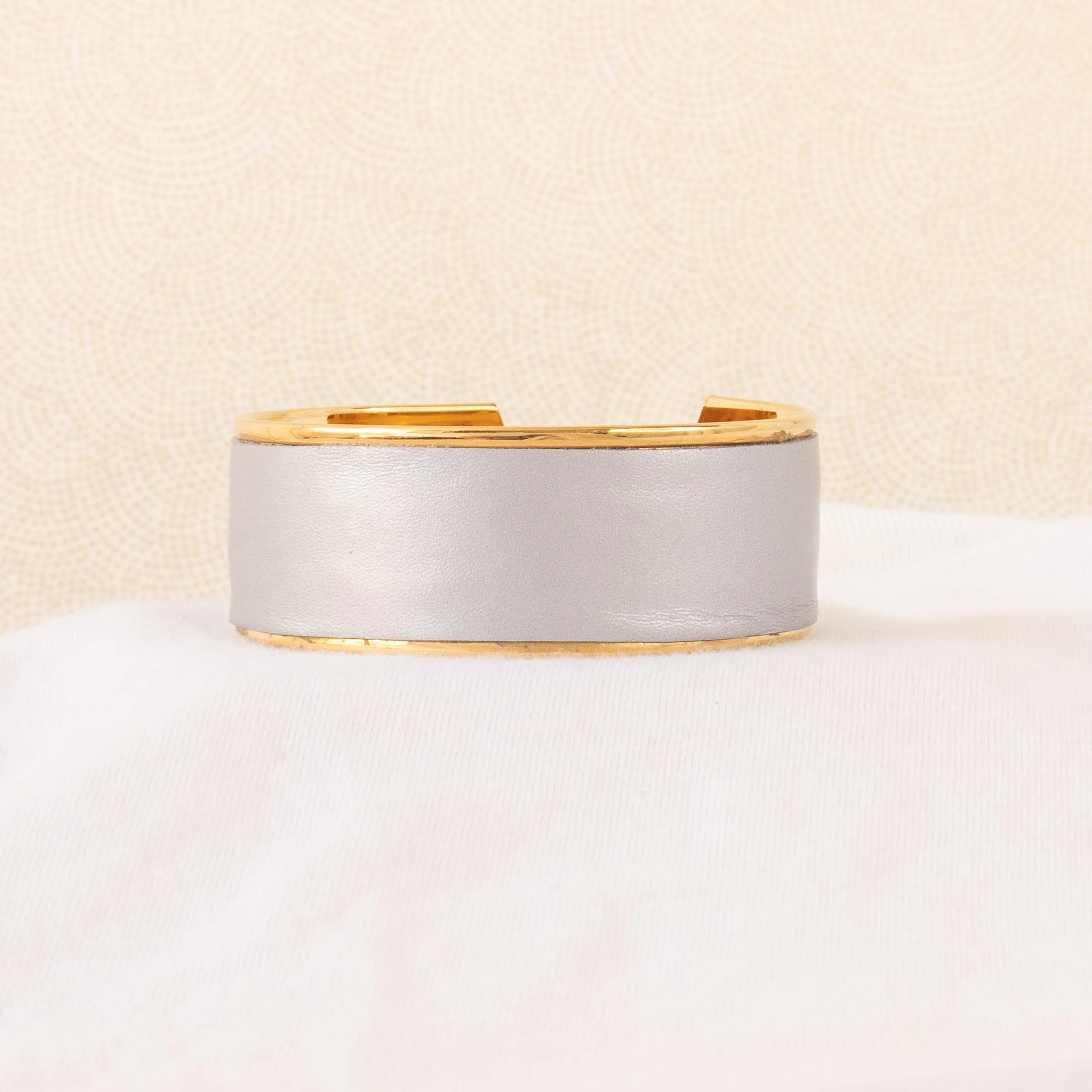 Bracelet en or et en cuir gris posée sur un tissu blanc avec un fond beige