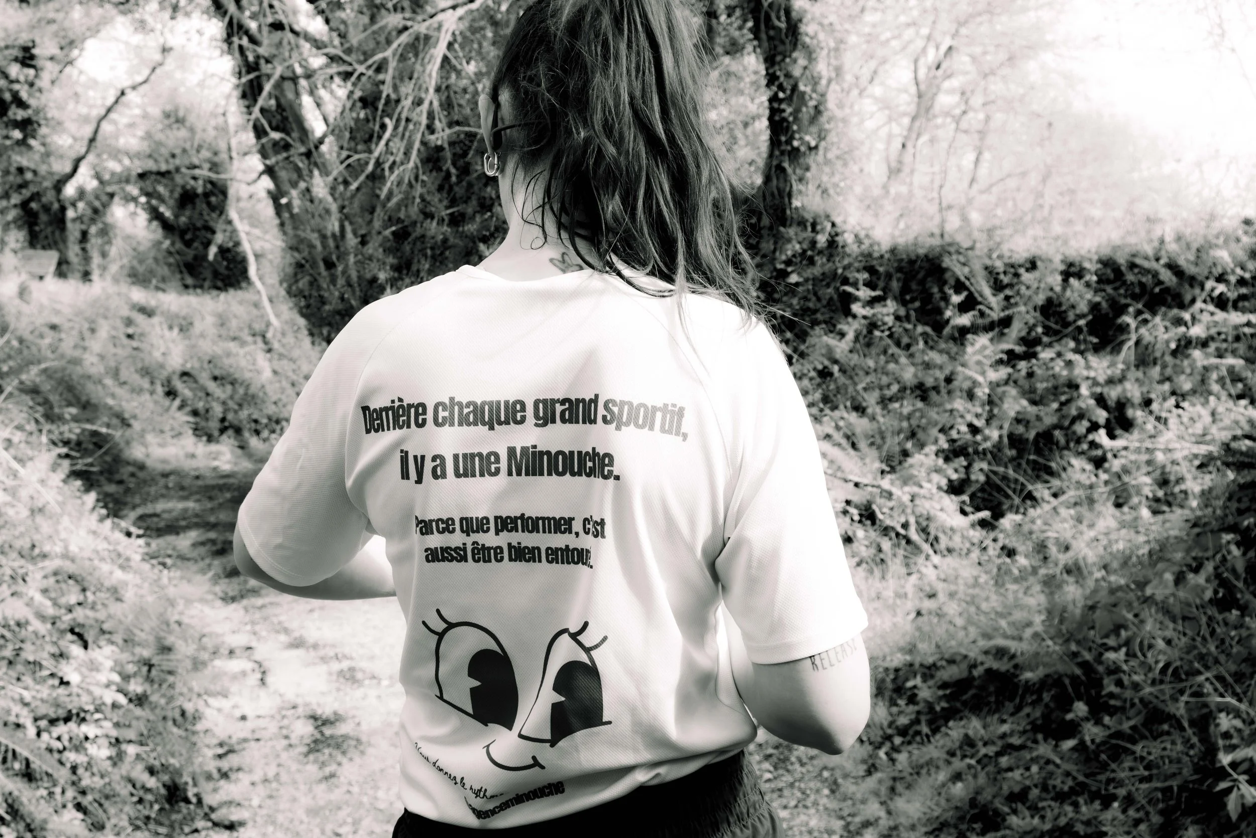 Une femme vue de dos, portant un t-shirt blanc avec un texte en français, dans une nature avec des arbres et un sentier.