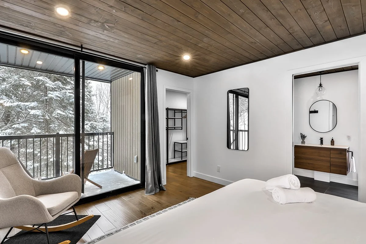 Chambre d'hôtel moderne avec un lit blanc, un canapé beige, un miroir mural, un balcon avec vue sur la neige, et un espace lavabo avec un miroir rond.