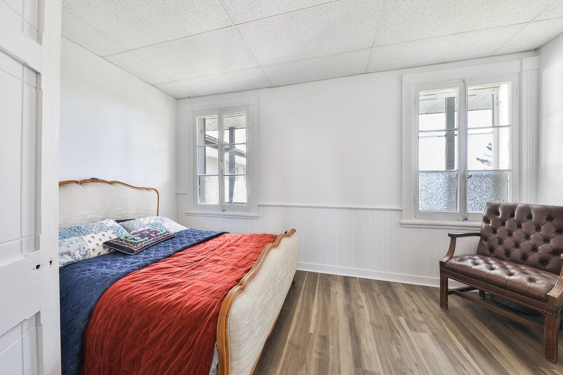 Chambre simple avec un lit ancien, oreillers colorés, et une banquette en cuir en bois, vue sur deux fenêtres avec vue extérieure, sol en bois, murs blancs et plafond suspendu.