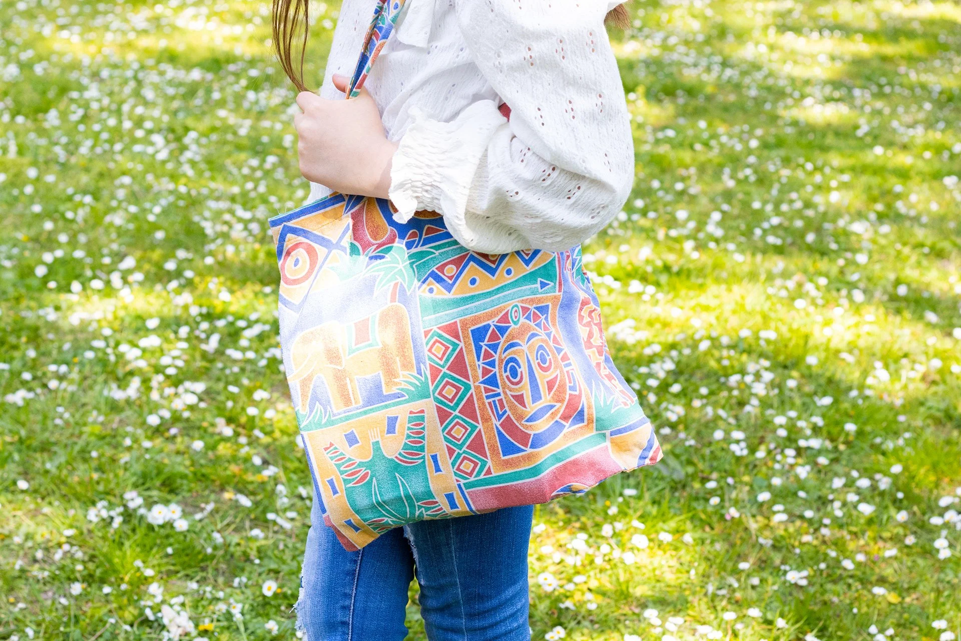 Une personne tenant un sac en tissu coloré avec un motif abstrait, dans un parc avec de la verdure et des fleurs blanches