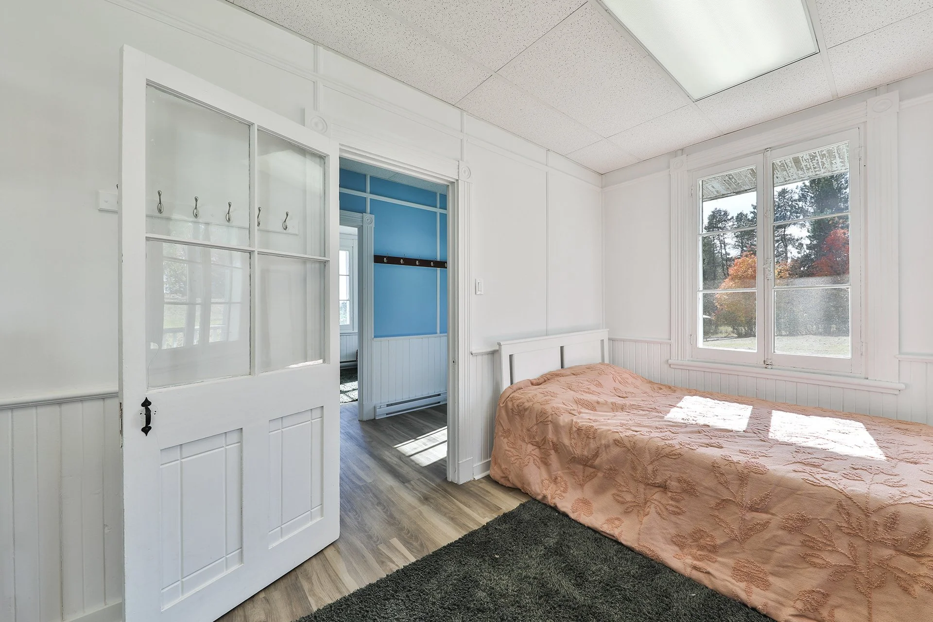 Chambre avec lit simple, fenêtre laissant passer la lumière, porte en bois blanc, sol en bois, mur blanc, pièce lumineuse.