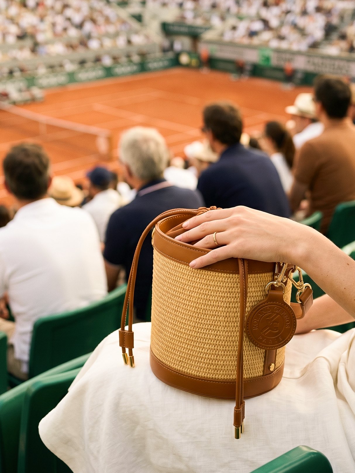 Une main tenant un sac beige et marron posé sur les genoux d'une personne assise dans un stade de tennis, avec le terrain en terre battue en arrière-plan et des spectateurs.