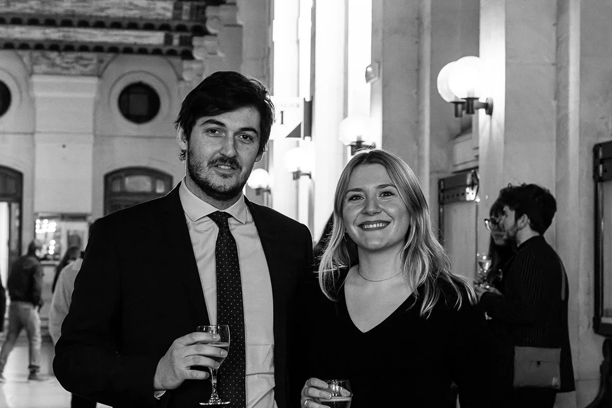 Deux personnes souriantes tenant des verres à vin dans une salle élégante avec des lumières murales, probablement lors d'une réception ou d'une fête.