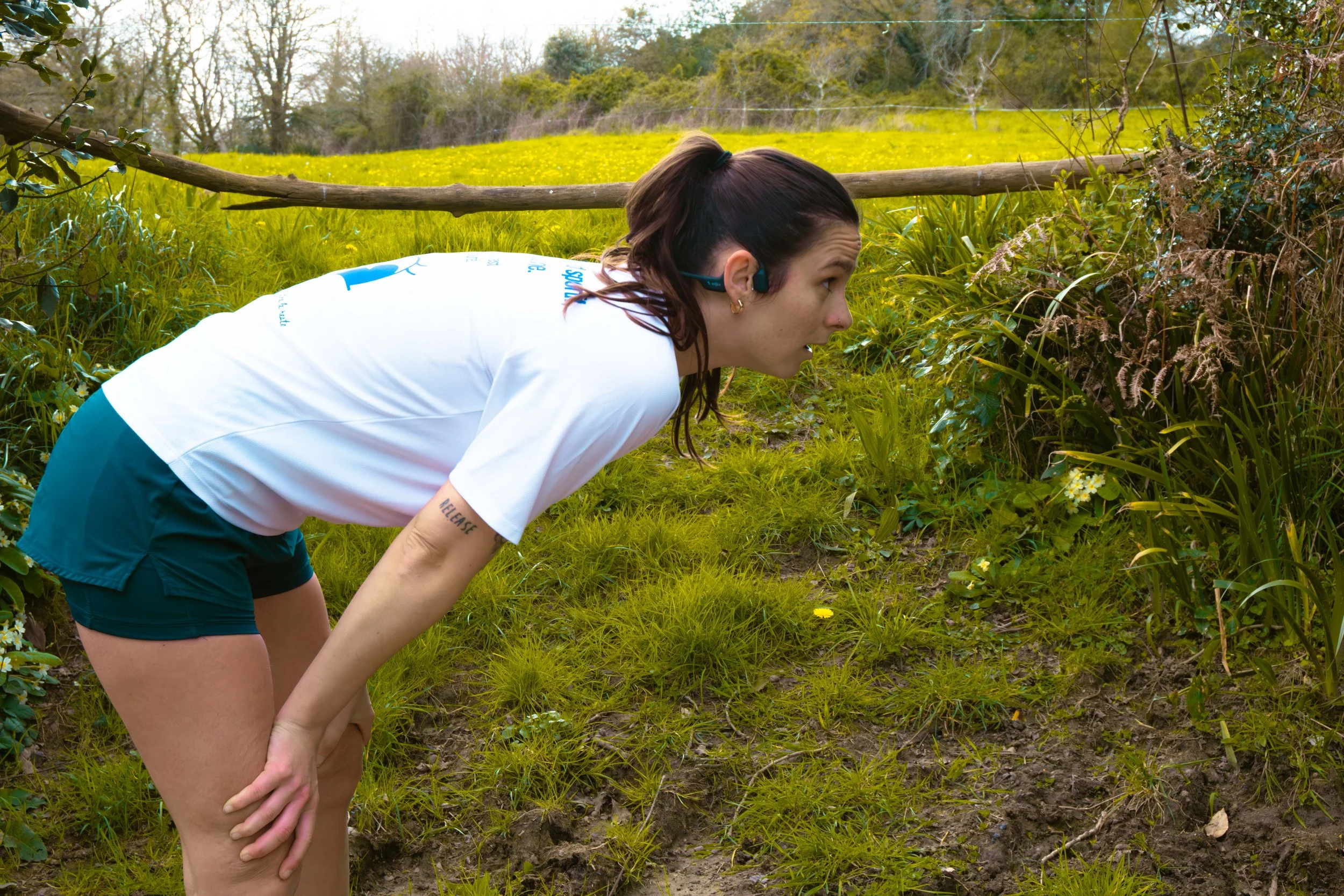 Femme en vêtement de sport, debout penchée en regardant près du sol dans une zone herbeuse en plein air.