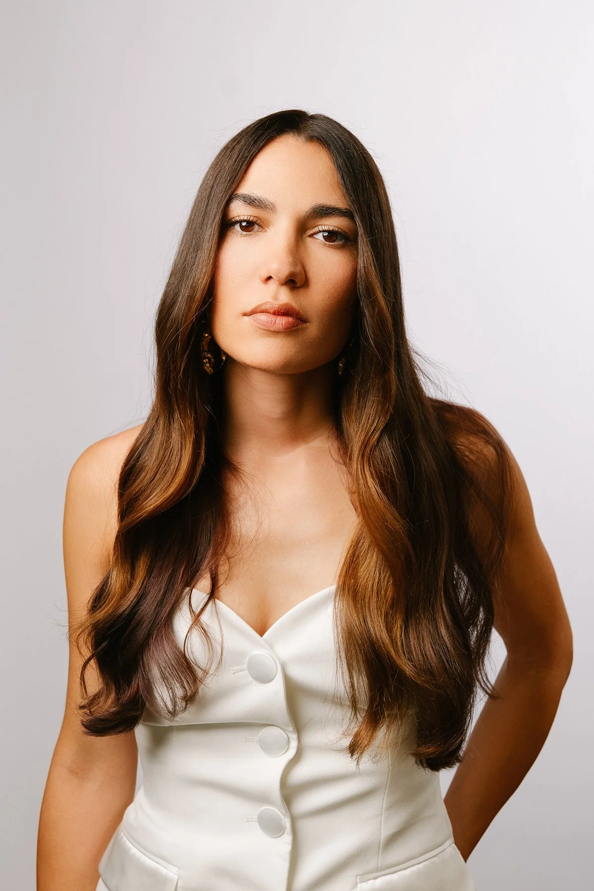 Portrait d'une femme avec de longs cheveux bruns, portant une robe blanche avec des boutons, face à la caméra contre un fond gris clair.