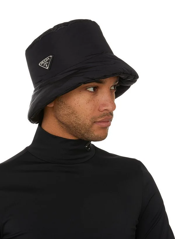 Un homme portant un chapeau noir en nylon avec le logo Prada, vêtement noir ajusté avec col montant.